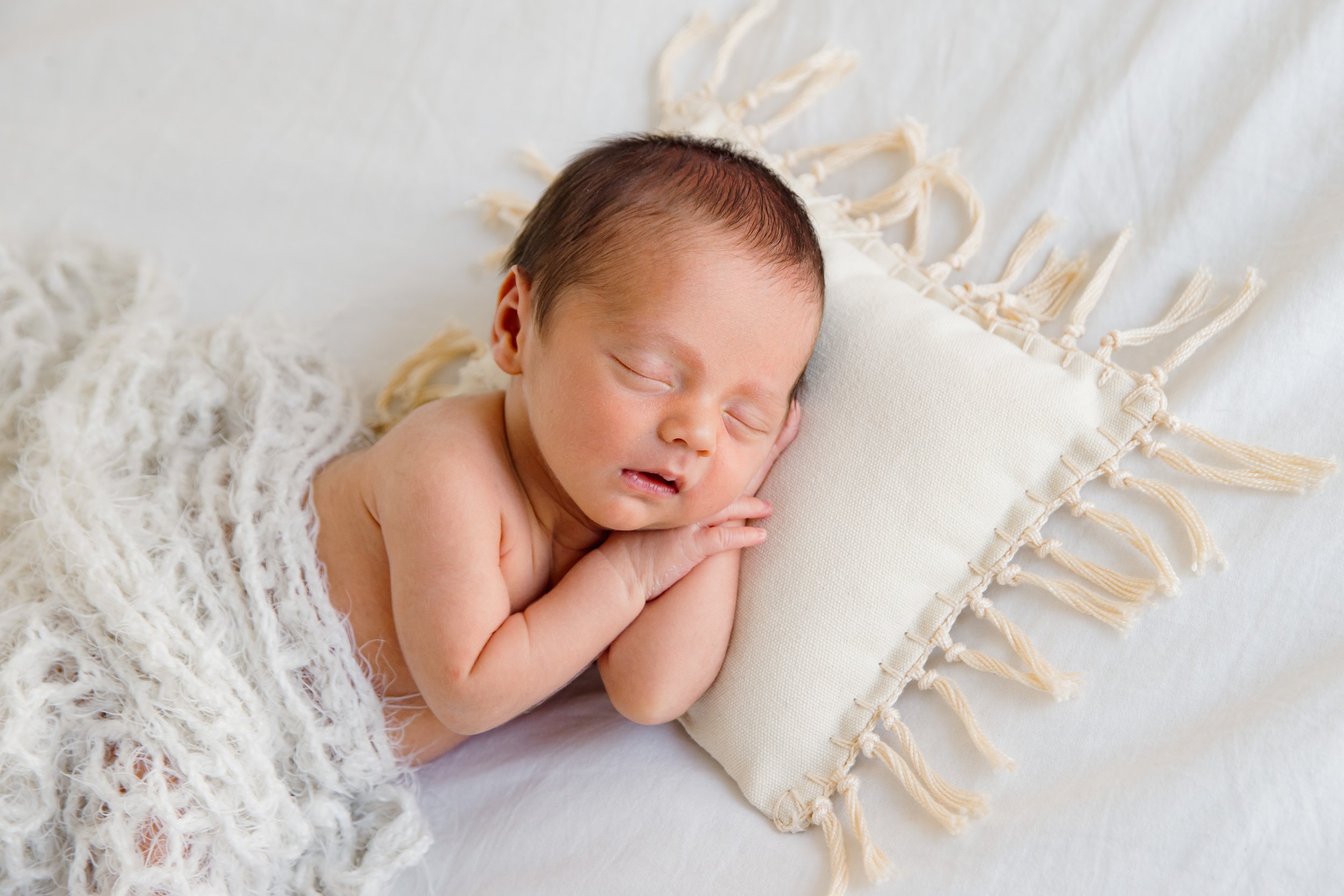 A newborn baby sleeping on a white blanket, resting on a decorative pillows with tassels.