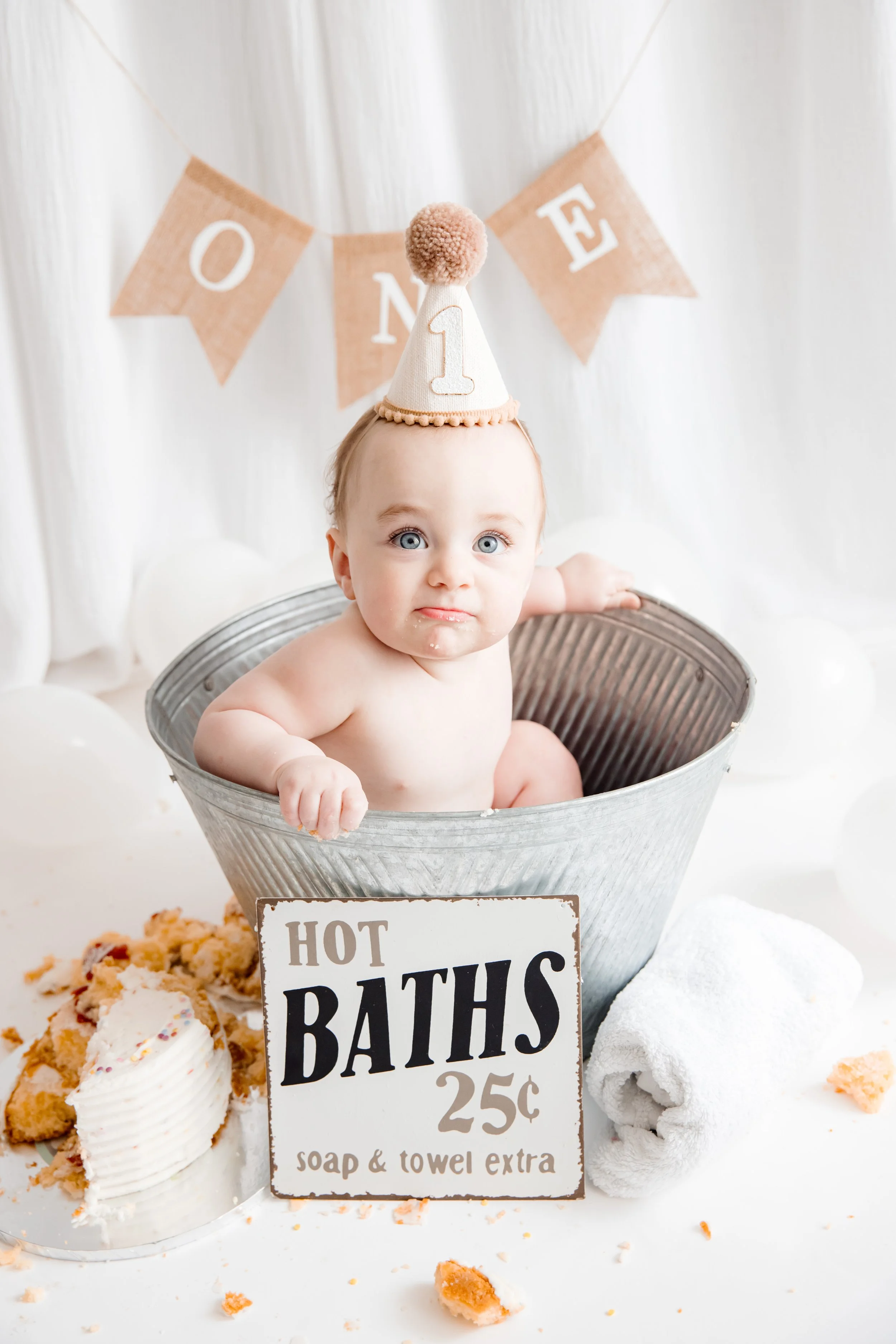 Young child with blue eyes and blonde hair wearing a birthday hat, sitting in a silver tub during a birthday celebration, with a "ONE" letter banner in the background and various desserts and a sign reading "Hot Baths 25¢ soap & towel extra" in front