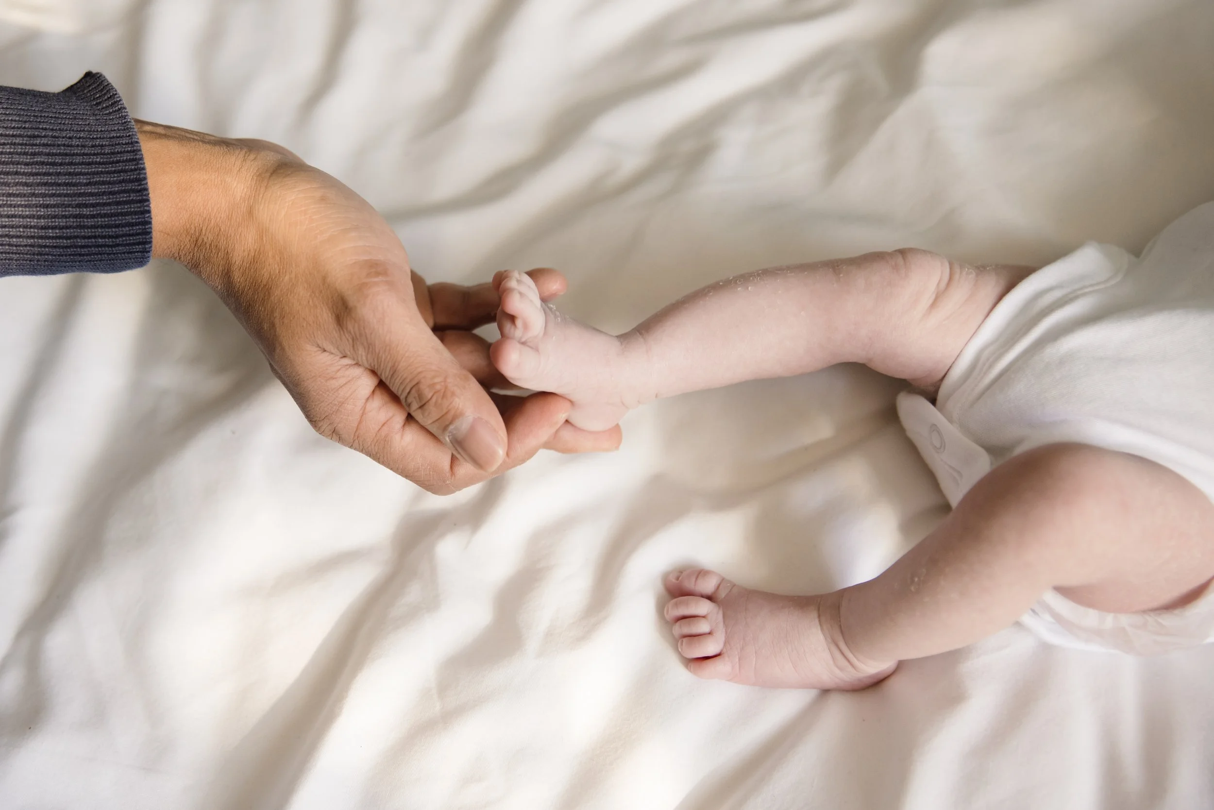 A close-up of an adult hand gently holding the tiny hand of a baby, with a neutral background and soft lighting.