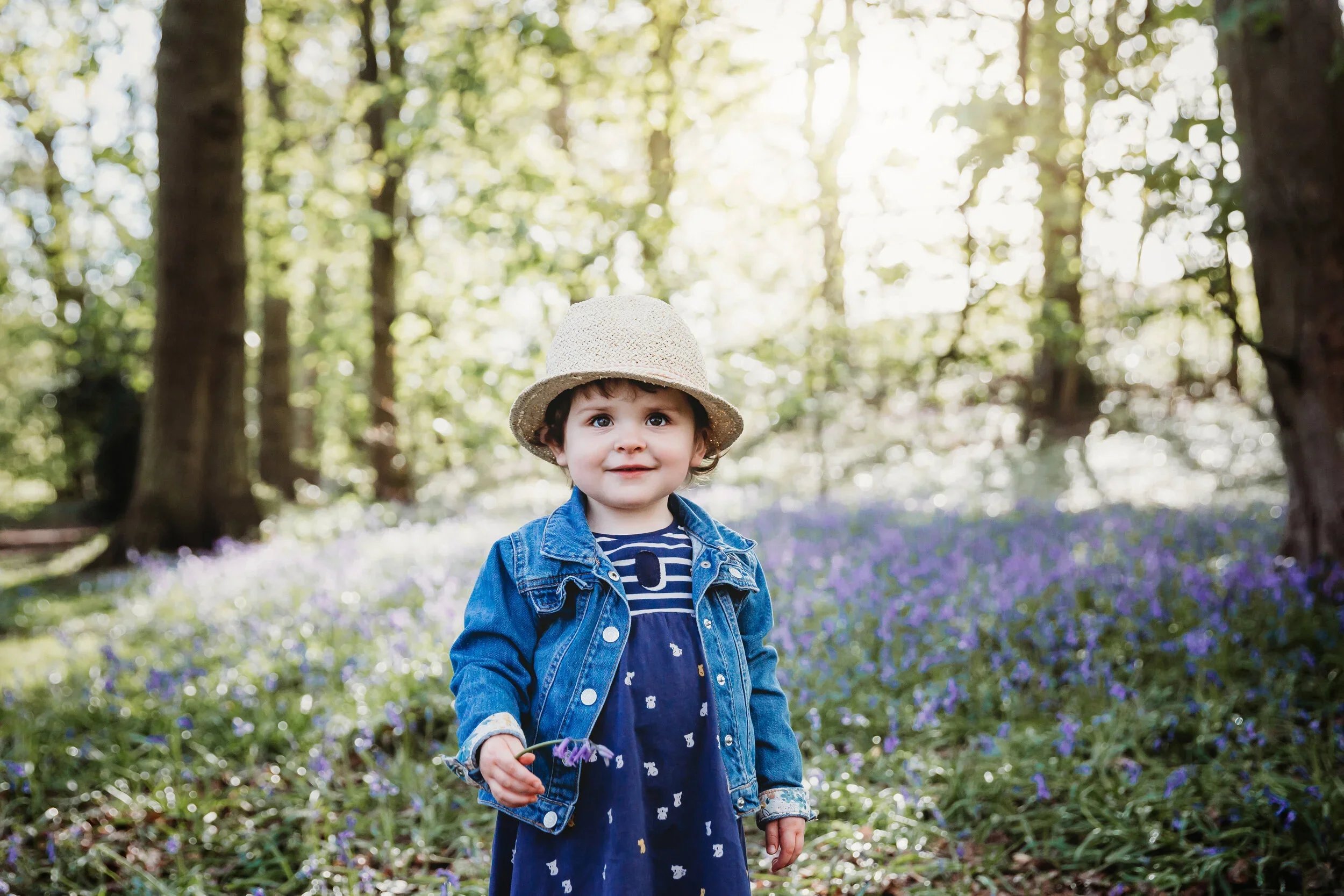 A young girl with curly hair wearing a straw hat, denim jacket, and a navy dress with white animal print, holding flowers, standing in a wooded area with sunlight filtering through trees and purple flowers on the ground.