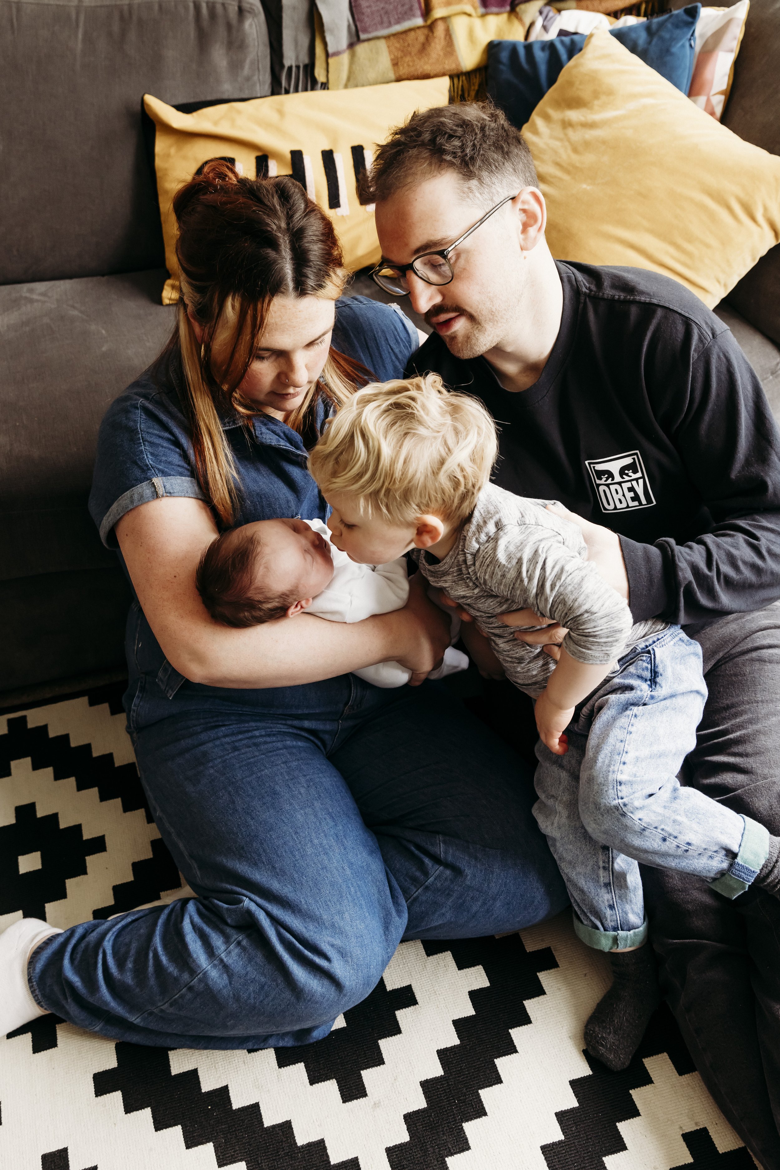 A family sitting on a black and white patterned rug in a room with a sofa and pillows in the background. A woman with red hair is holding a newborn baby, while a man with glasses and a young boy with blonde hair are looking at the baby. The family ap