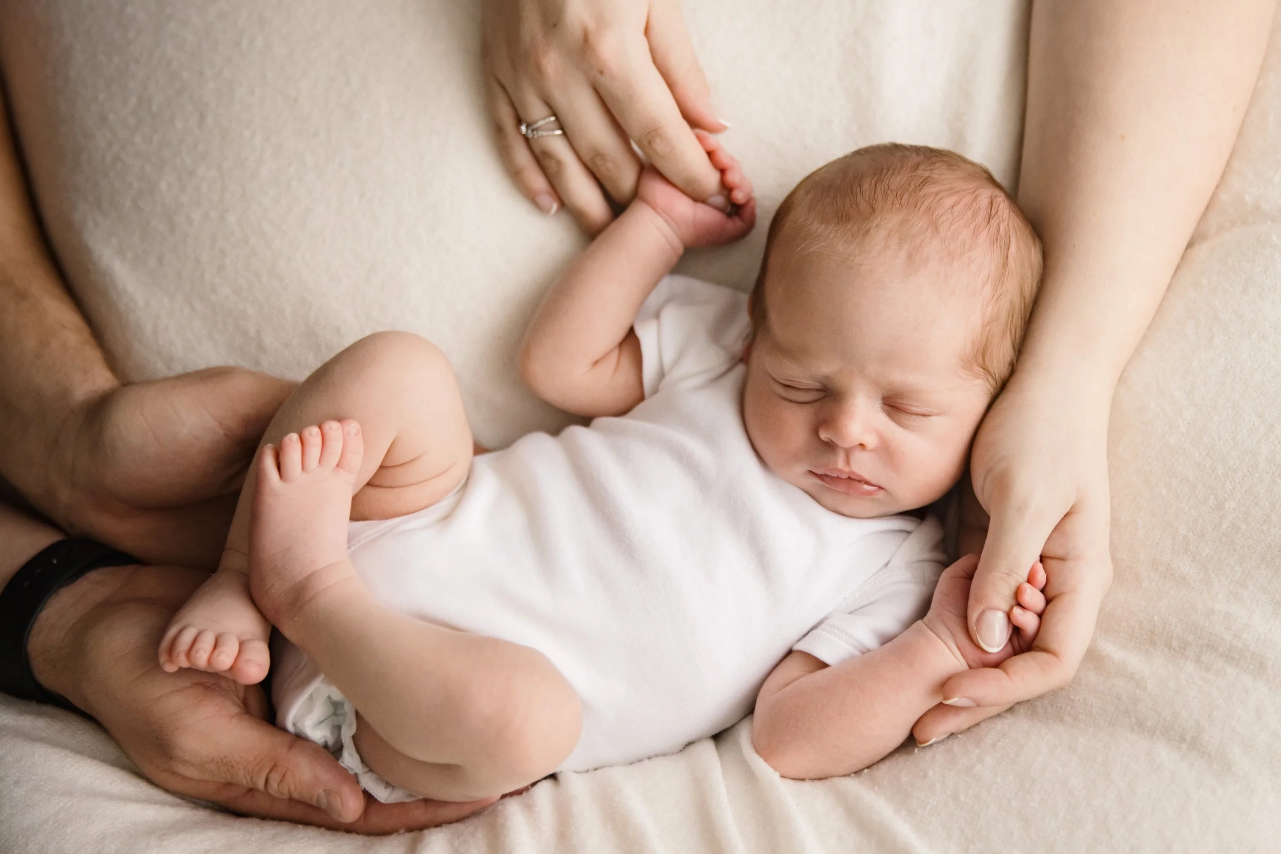 A newborn baby is sleeping on a soft surface, with parents holding the baby's small hands and feet gently. The baby's head is resting on the parent's arm, and the parent is wearing a wedding ring.