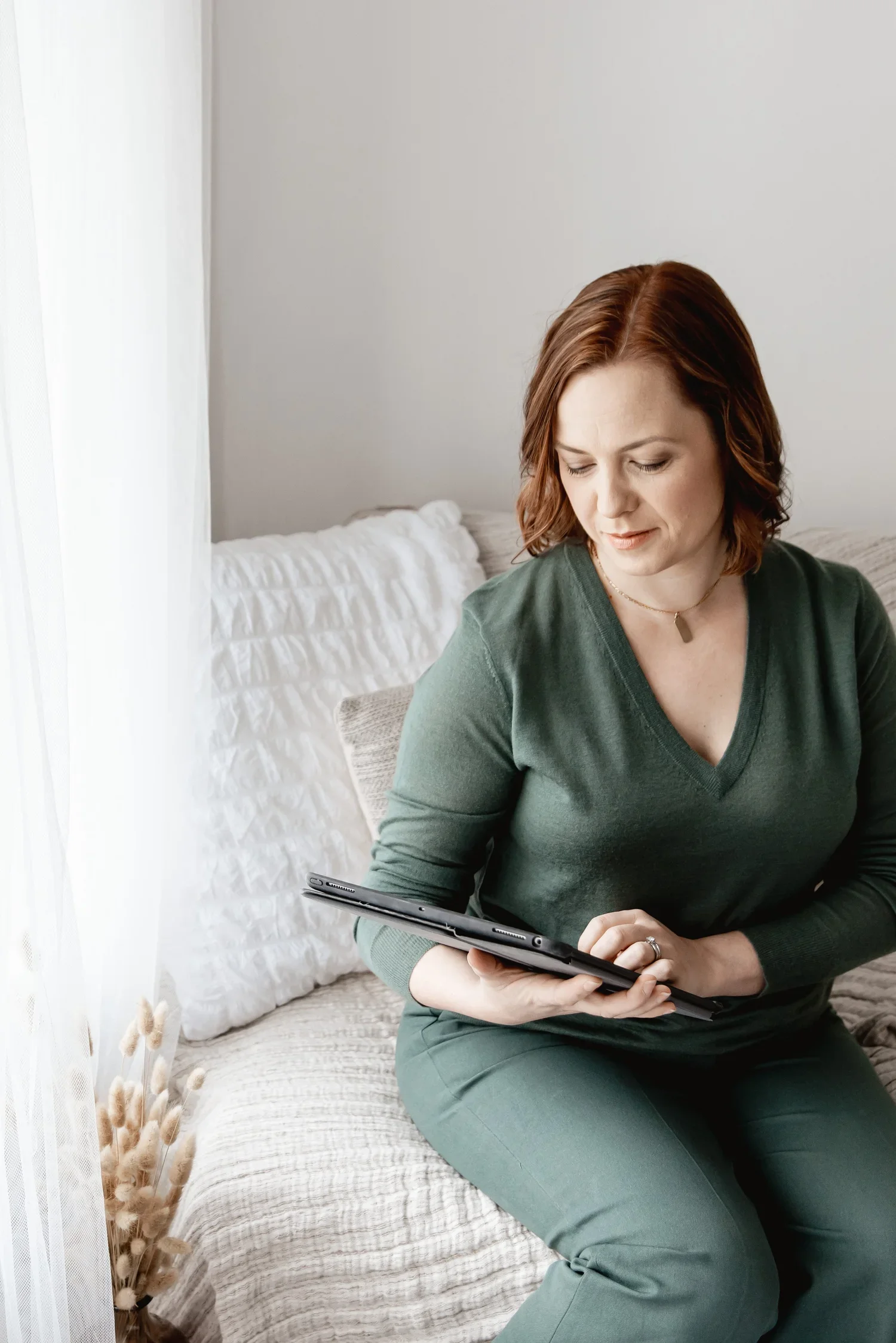 A woman with brown hair, wearing a green long-sleeve top and green pants, sitting on a bed and looking at a tablet in her hands.