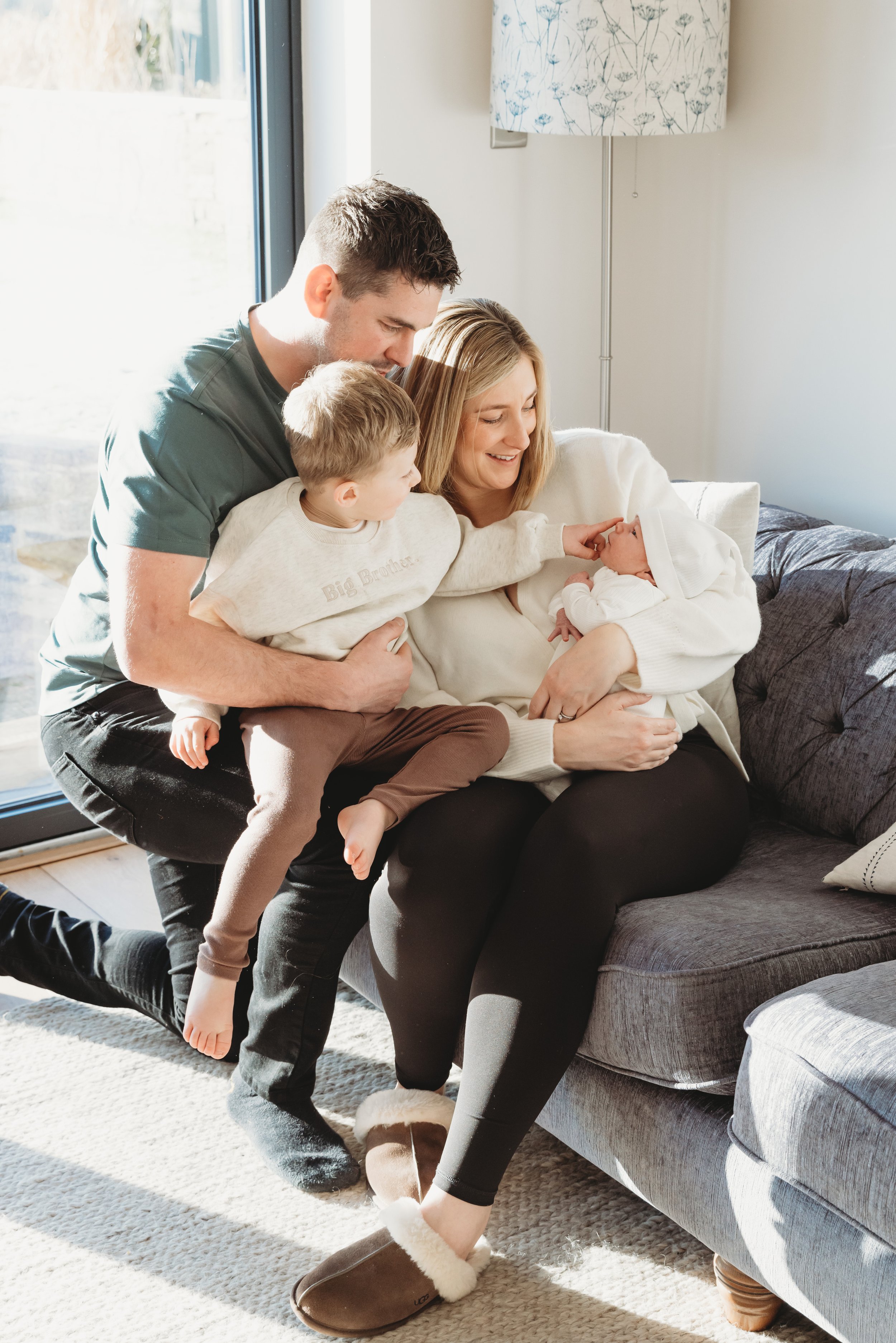 A family of four sitting on a gray couch in a living room, with sunlight coming in through a large window. The mother is holding an infant dressed in white, and their young son is touching the baby's face while standing close. The father is kneeling 