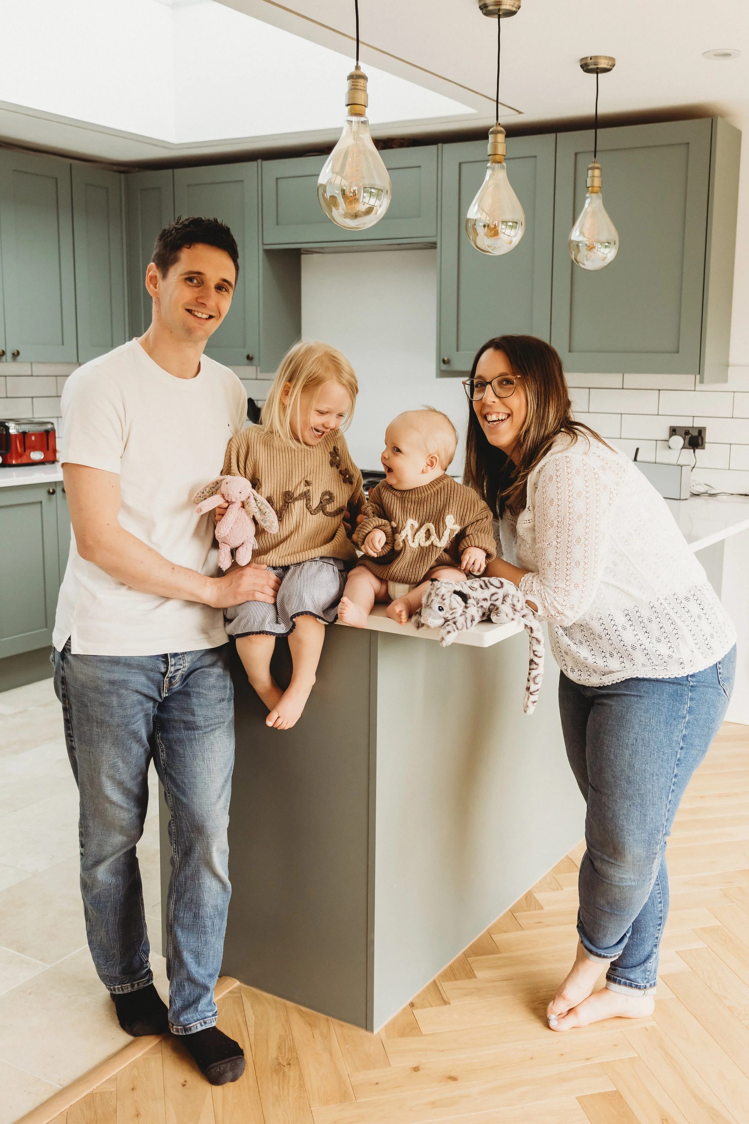 A smiling family of four in a modern kitchen. The father is wearing a white t-shirt and jeans, holding a plush bunny, with his feet on the floor. The mother is wearing glasses, a white lace top, and jeans, while barefoot, leaning on the kitchen islan