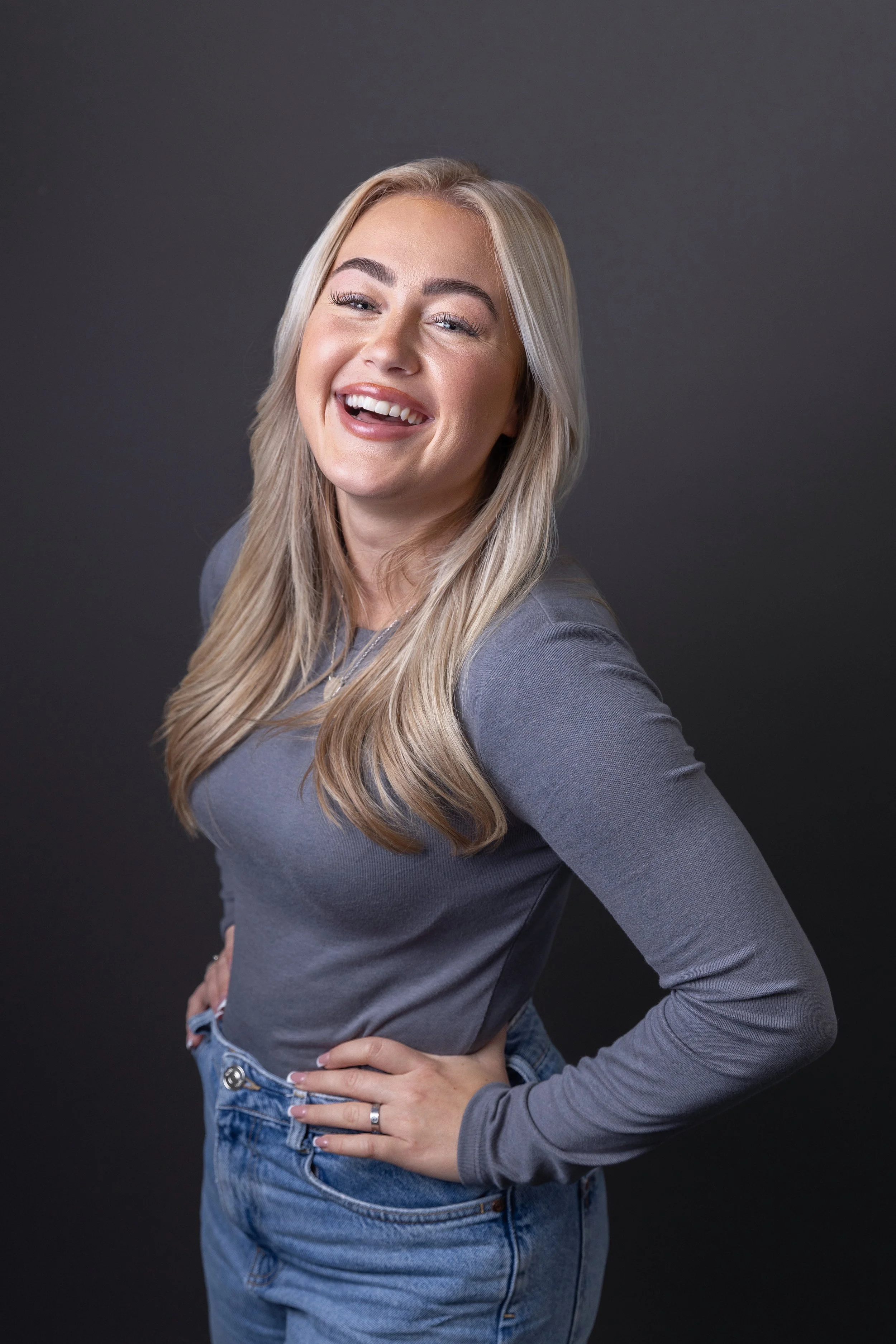 A woman with long blonde hair winking and smiling, standing against a dark gray background, wearing a gray long-sleeve shirt and blue jeans.