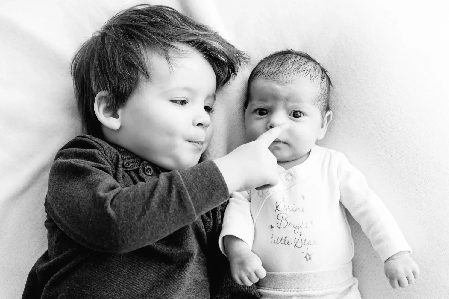 A young boy is gently touching the nose of an infant girl lying next to him, both lying on a soft surface.