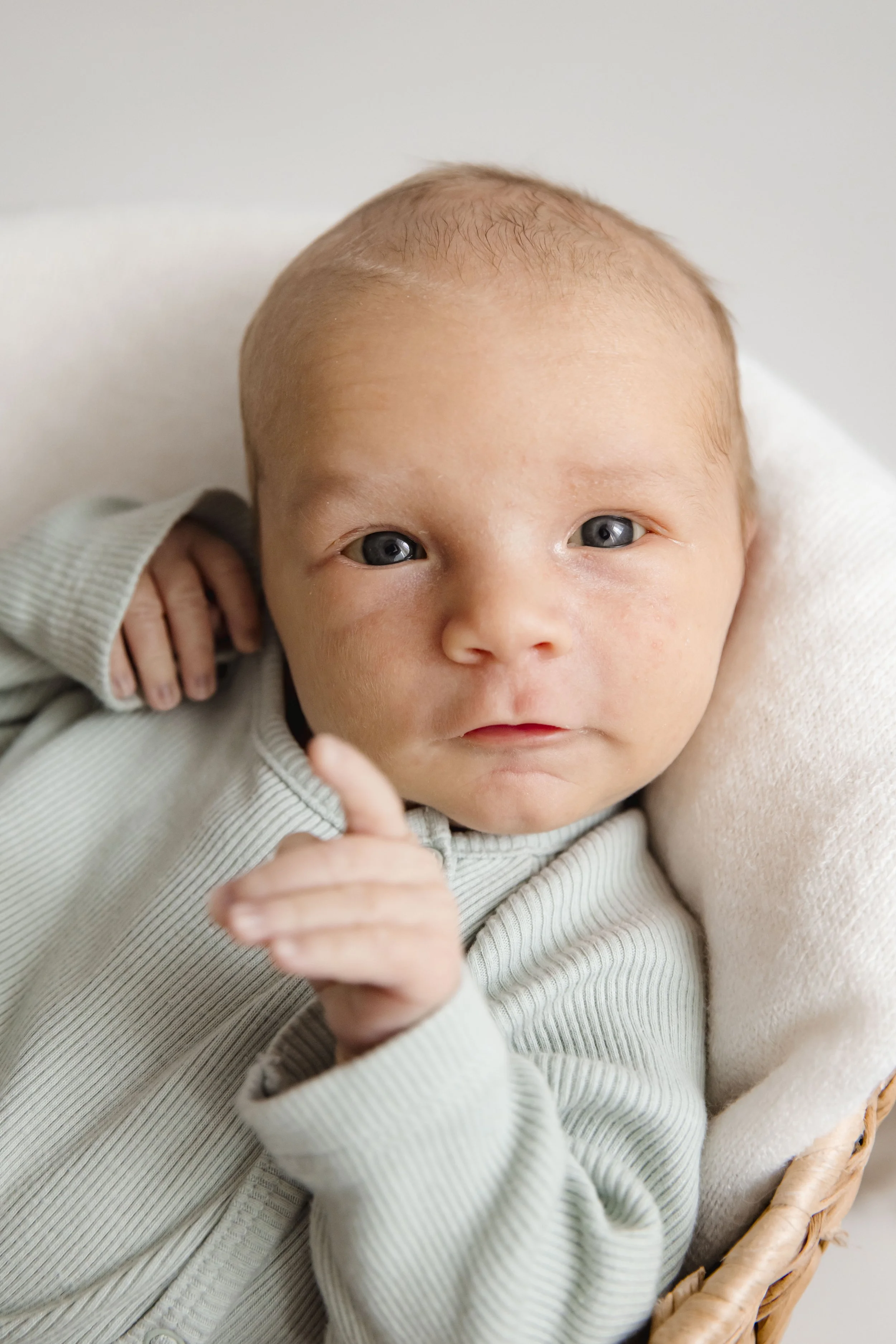 A close-up of a baby's face with blue eyes, lying in a basket, wearing a light-colored, ribbed long-sleeve shirt, with one hand near face and the other hand resting on the basket's edge.