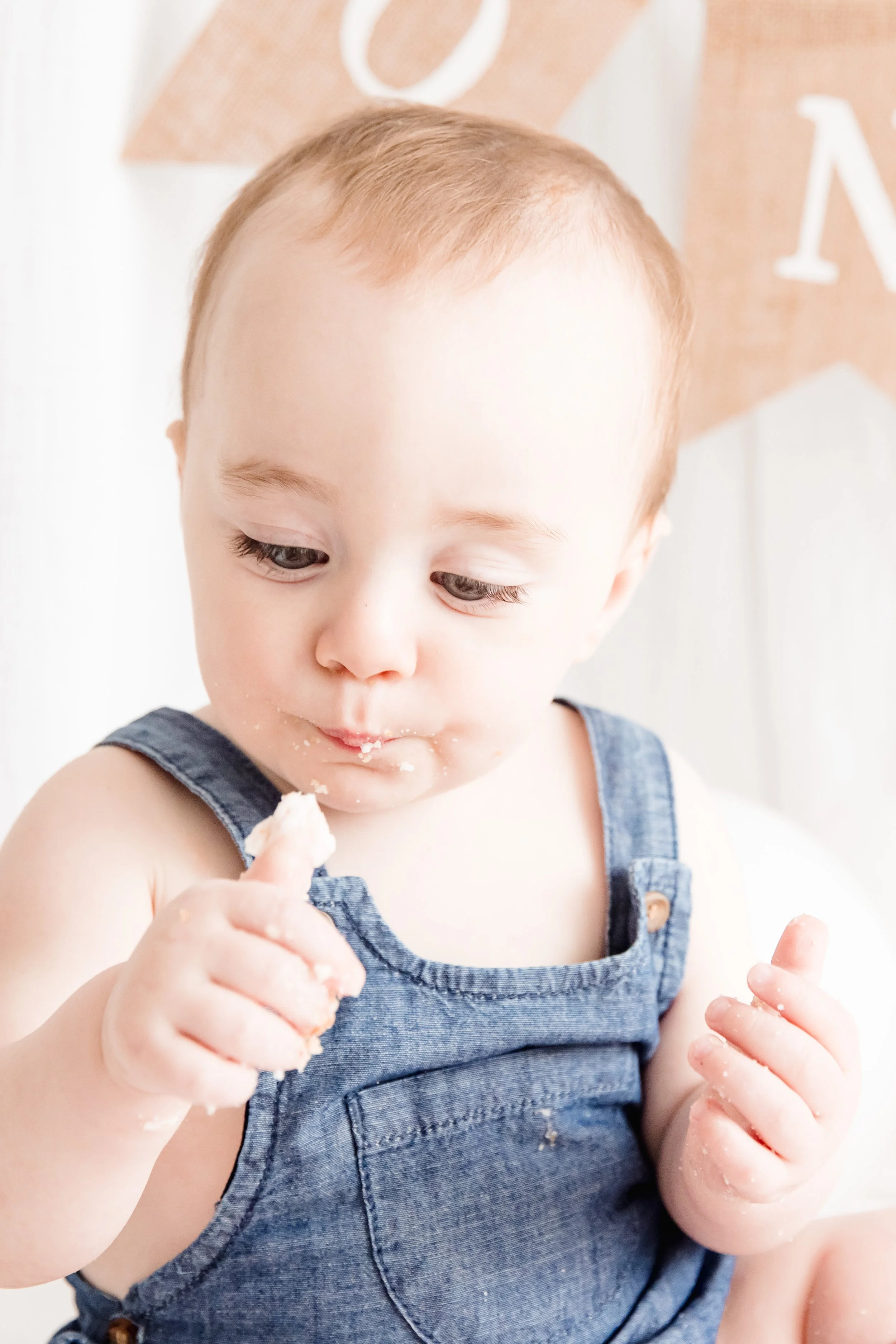A young child with light skin and short light brown hair, wearing a denim sleeveless top, is eating something with cream or frosting on their face and hands.