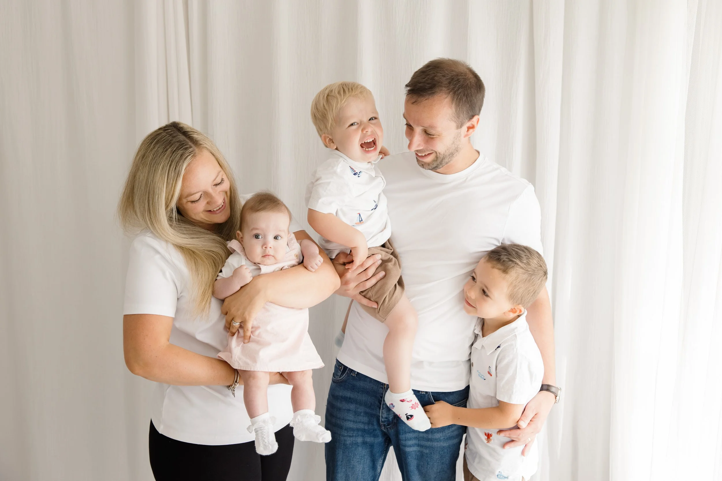 A family of six smiling and laughing together, including two women, a man, and three young children, standing against a white curtain background.