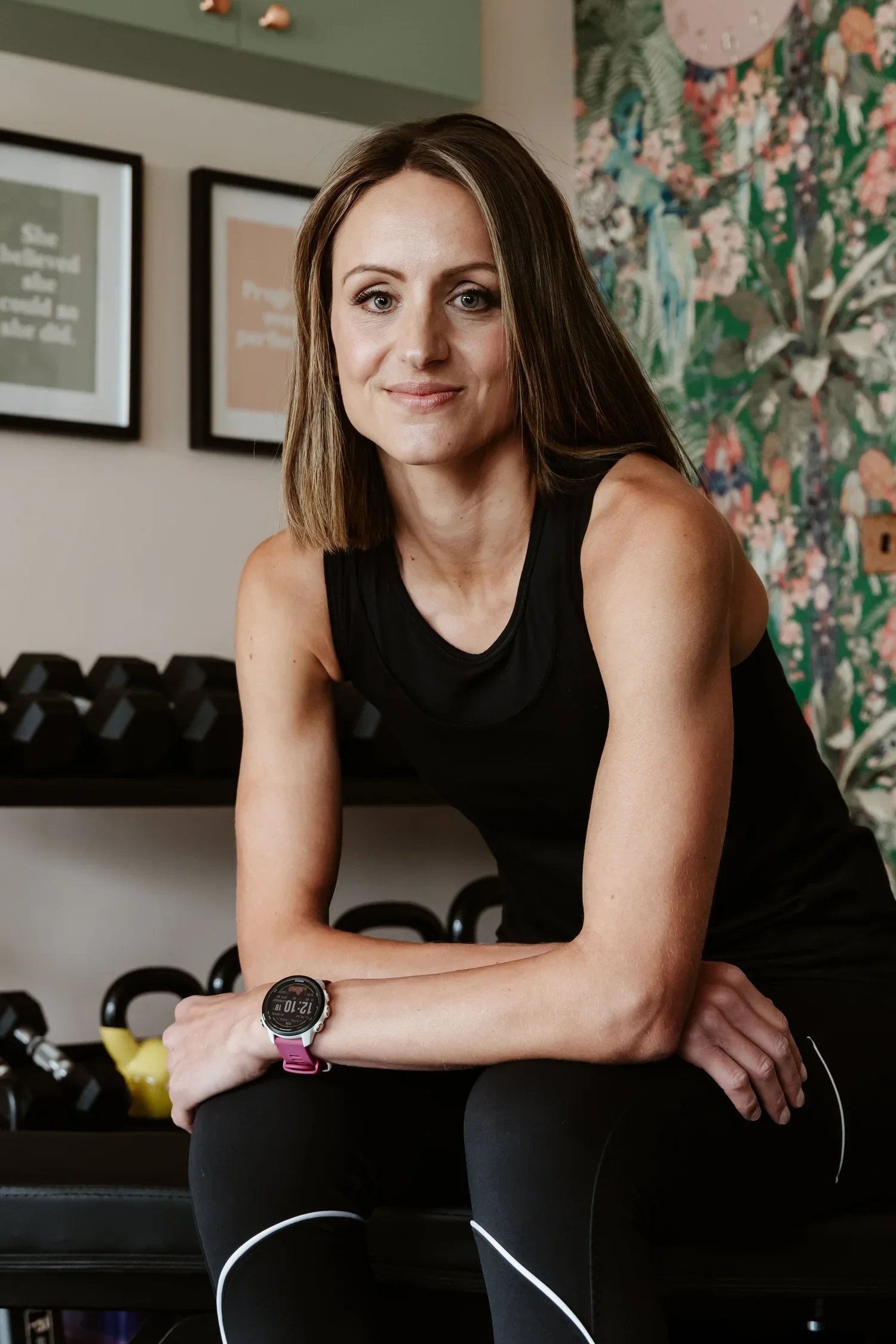 A woman with shoulder-length brown hair and light skin, wearing a black sleeveless top, sitting in a fitness studio with a wristwatch, surrounded by gym equipment and colorful wall art.
