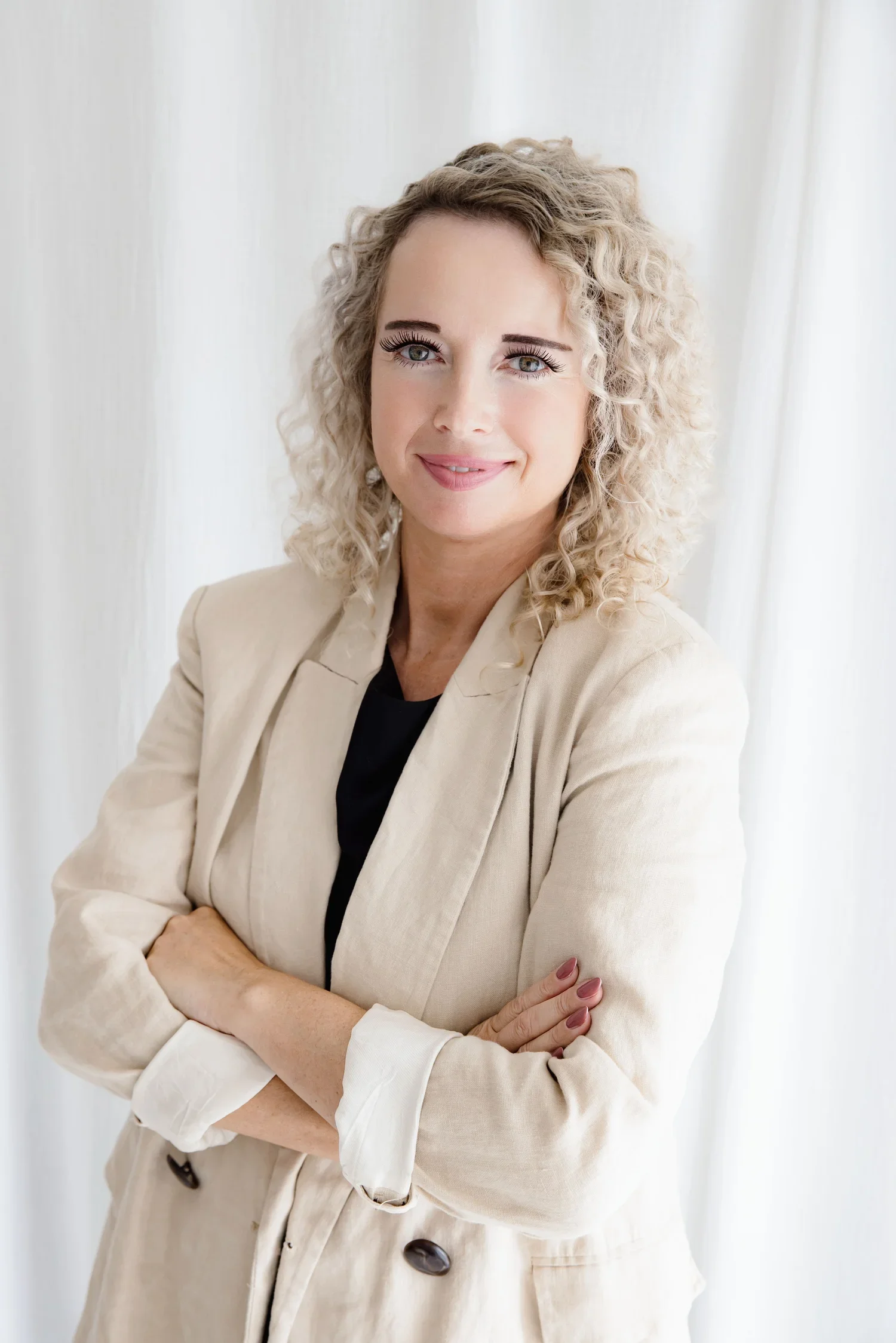 A woman with curly blonde hair smiling and crossing her arms, wearing a beige blazer and a black top, against a light background.