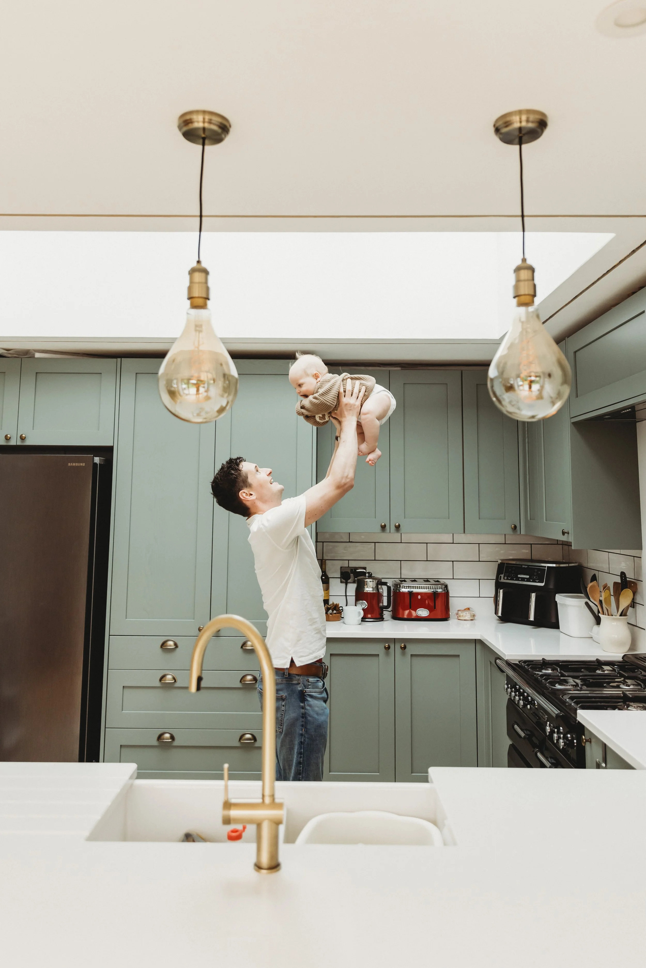 A man lifting a baby in a kitchen with green cabinets and pendant lights.