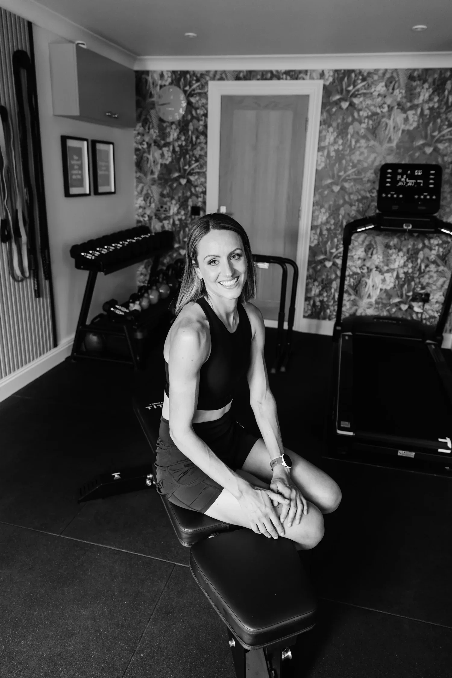 A woman sitting on an exercise bench in a home gym. She is smiling, wearing a sports bra and shorts, with workout equipment and floral wallpaper in the background.