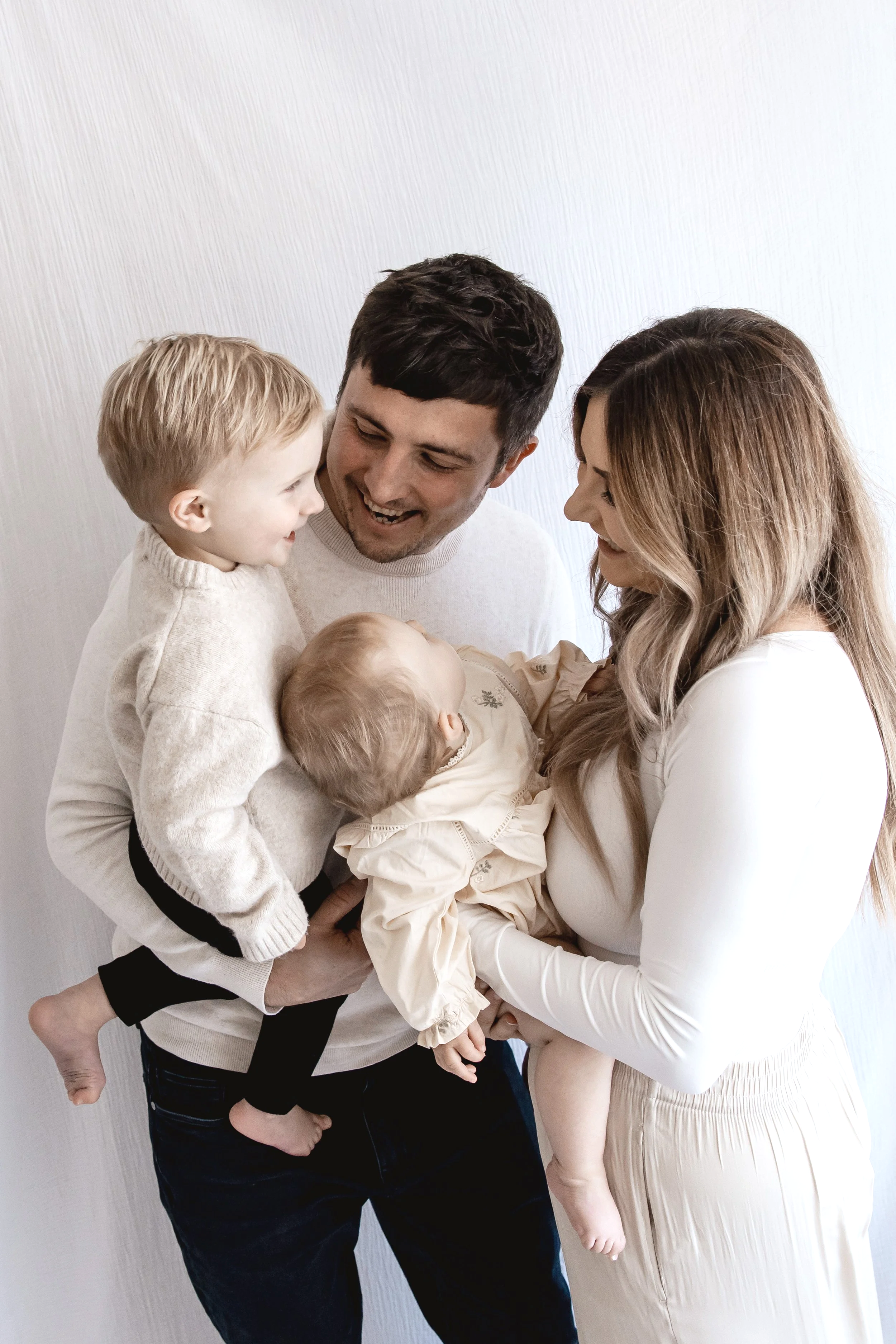A happy family of five celebrating, with a father holding a young girl, mother holding a baby, and another girl standing close, all smiling and engaging warmly.