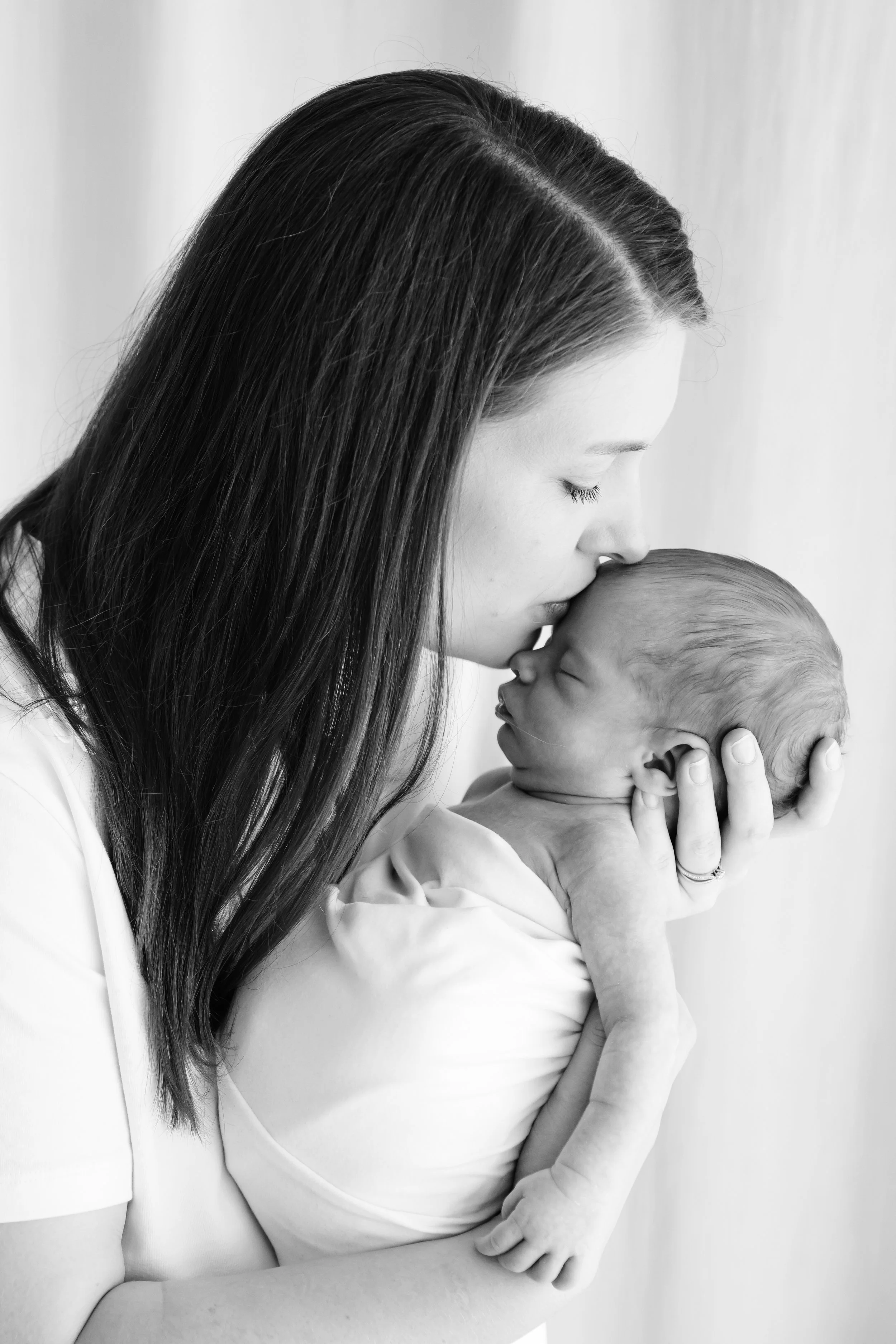 A woman with long hair gently holding and kissing the forehead of a baby, both in a close-up black and white photo with a plain background.