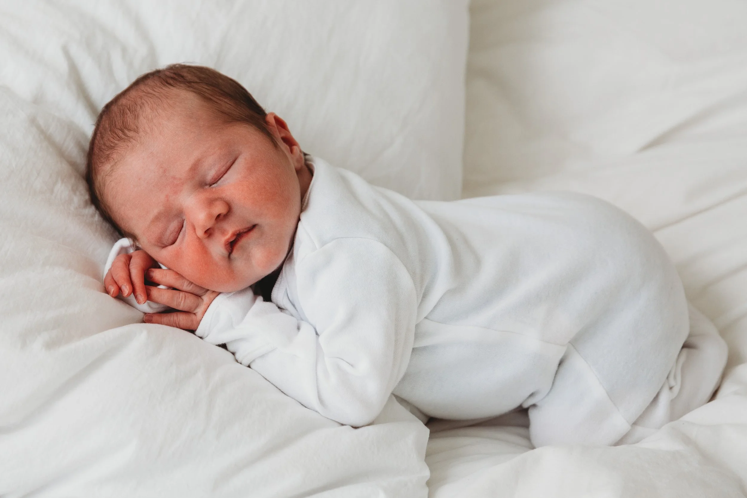 Close-up of a sleeping newborn baby with reddish-brown hair, dressed in white, resting on a soft white blanket.