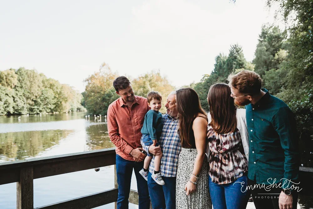 Family of six standing on a lakeside dock, smiling and talking, in a lush green outdoor setting.