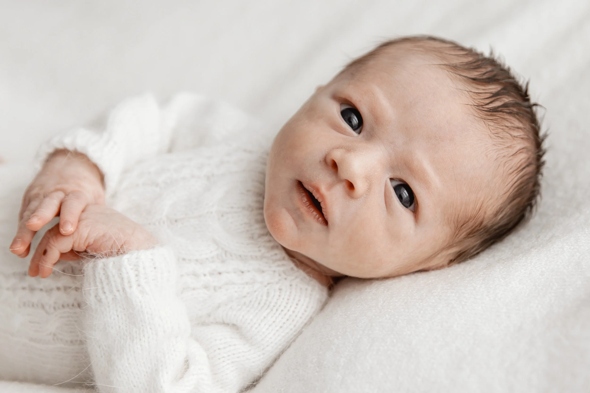 Close-up of a baby with big eyes lying on a white soft surface, wearing a white knitted sweater.