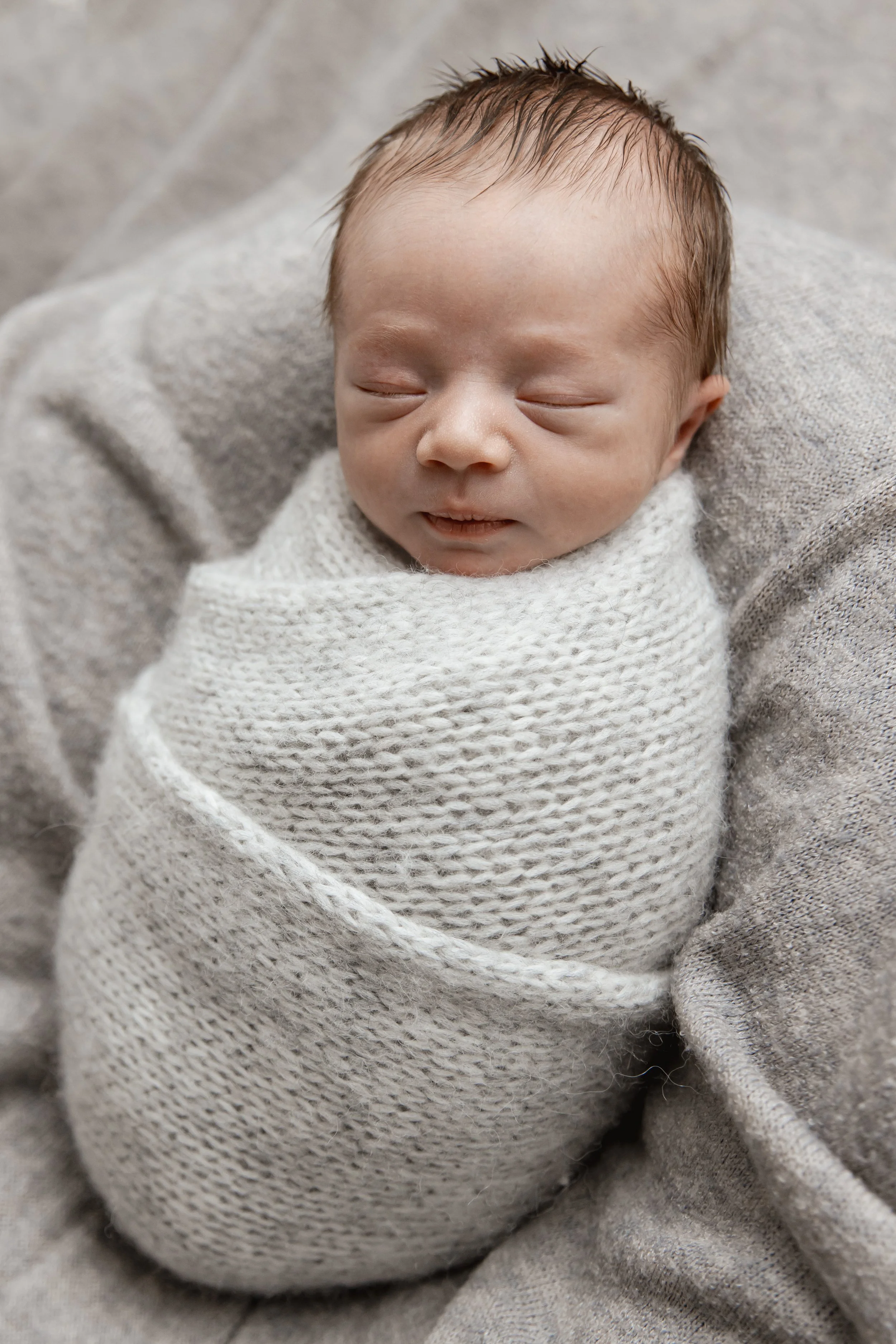 Close-up of a sleeping newborn baby wrapped in a white knitted blanket, lying on a soft gray fabric.