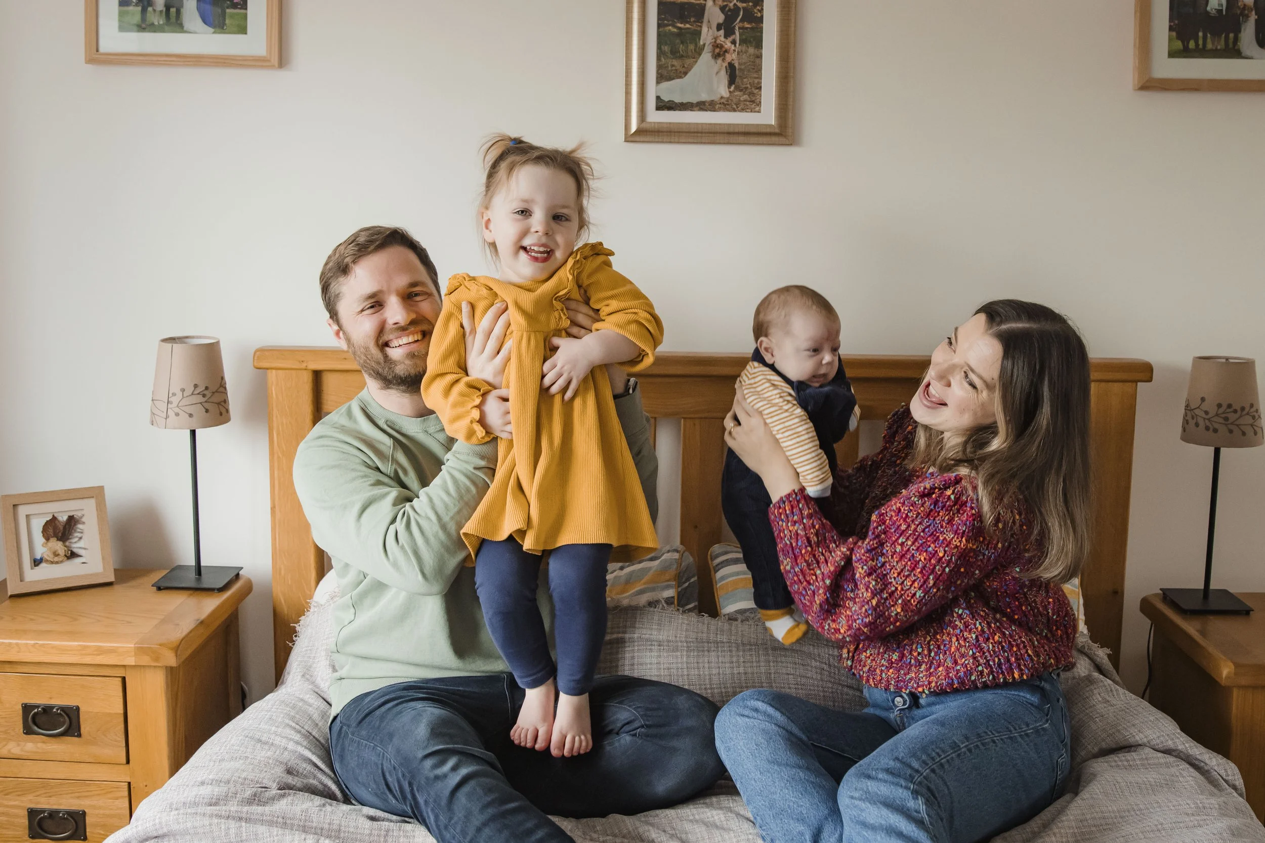 A family of four, including a father, mother, and two young children, smiling and enjoying time together in a living room. The father is sitting on a couch holding a girl in a mustard yellow dress, and the mother is holding a baby boy. The background