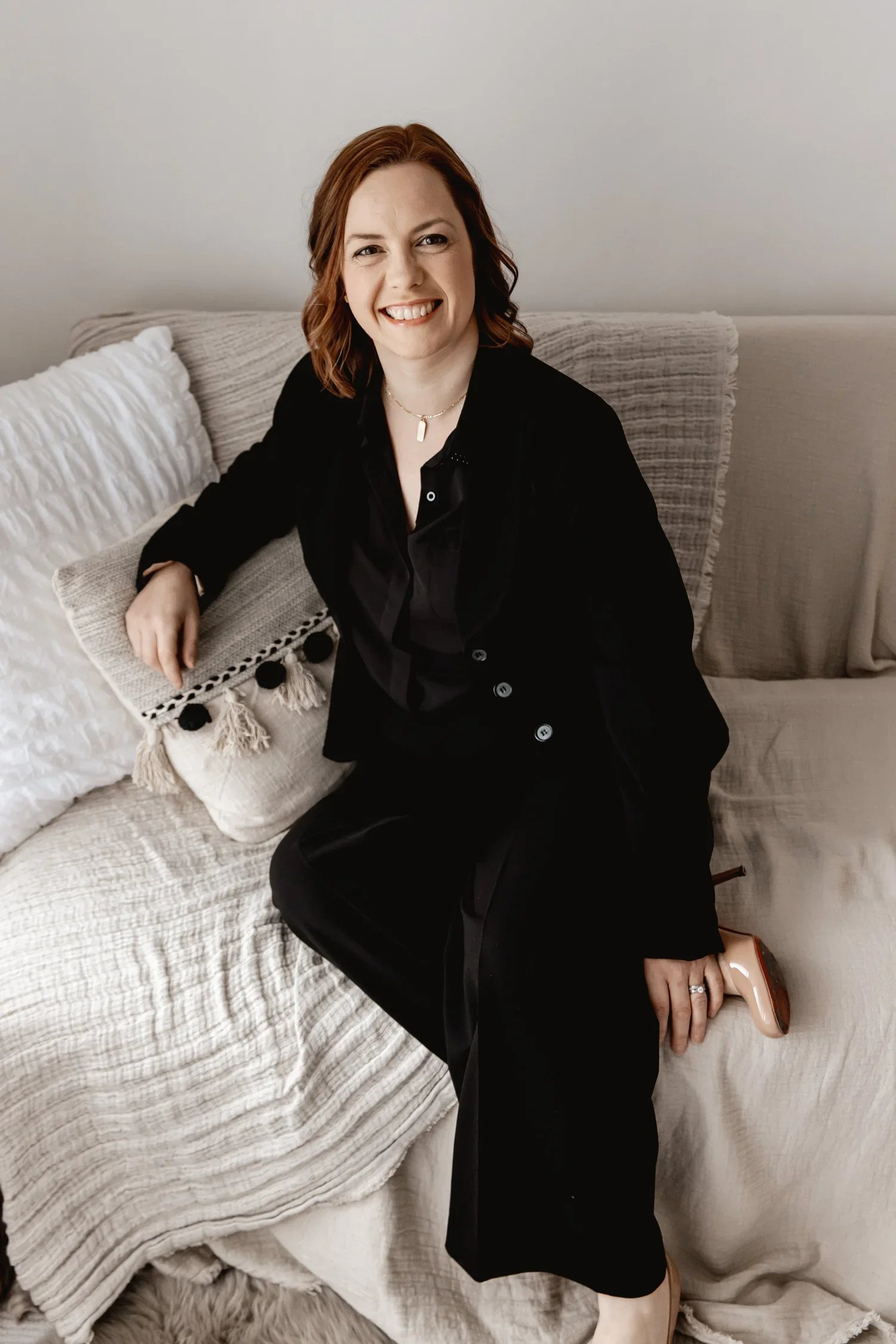 A woman with shoulder-length red hair smiling and sitting on a beige couch, wearing a black outfit and peach-colored heels, with a white cushion beside her and a light-colored blanket on the sofa.