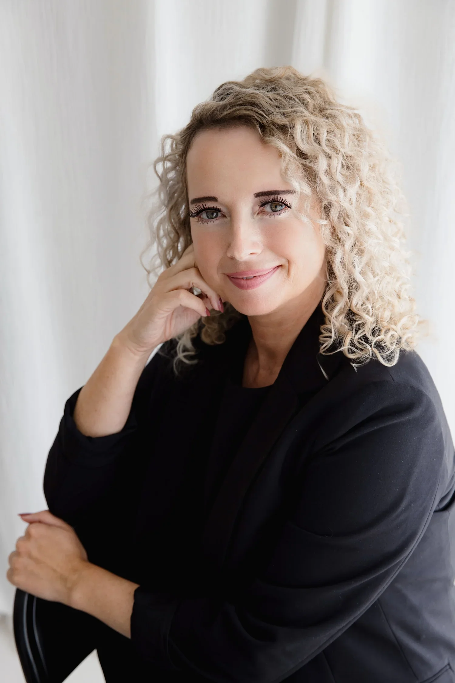 Headshot of a woman with curly blonde hair, wearing a black top, sitting with her hand near her face, smiling softly at the camera against a light background.