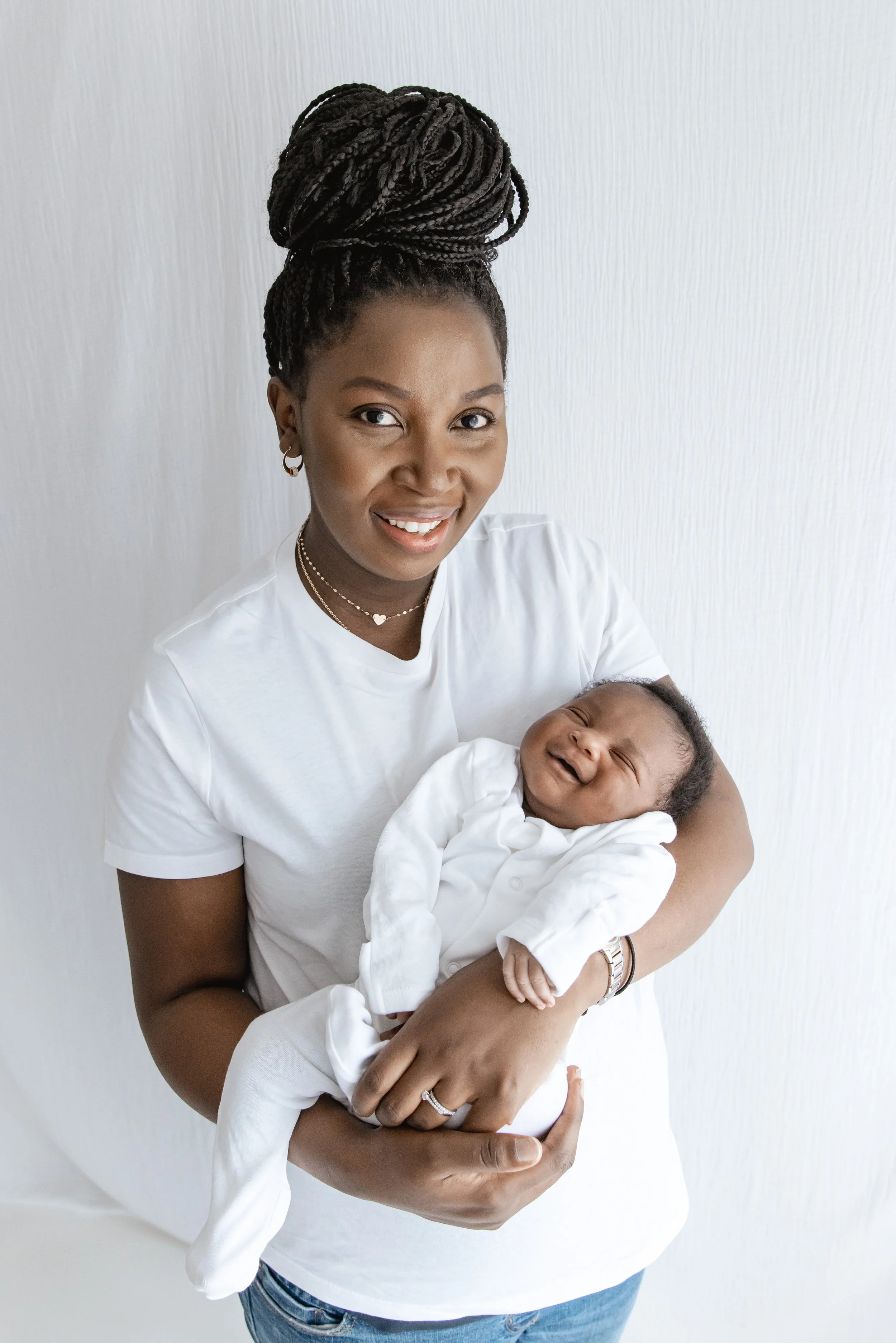 A woman with braided hair styled in a high bun holding a laughing baby in her arms against a plain white background.