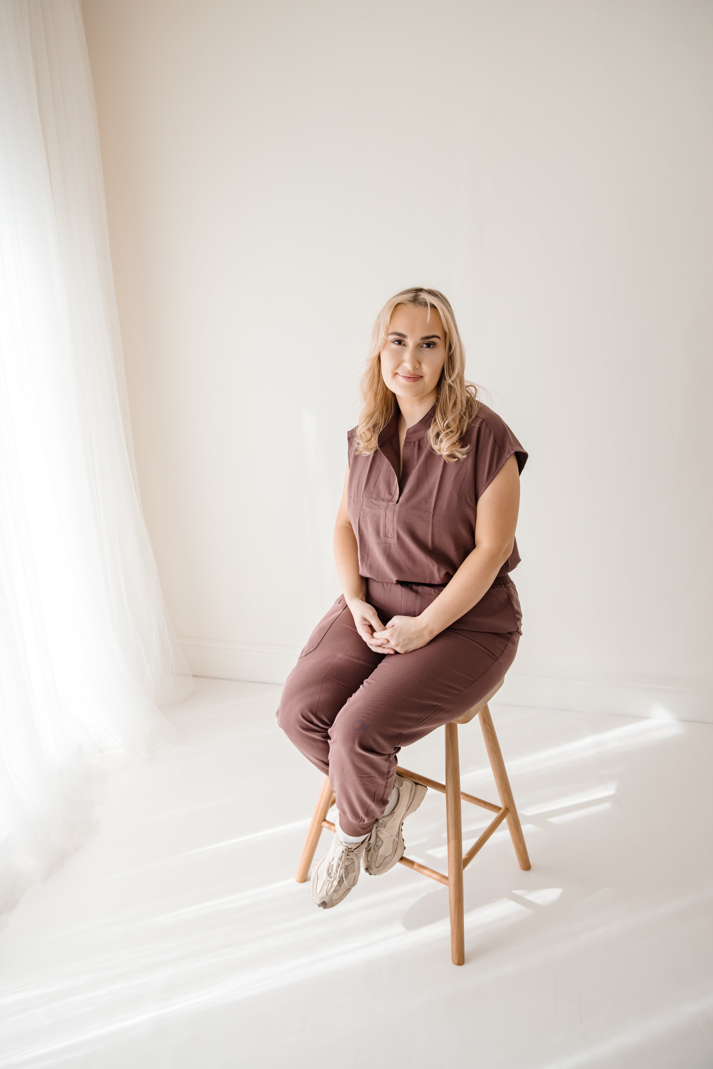A woman with blonde hair sitting on a wooden stool in a minimalistic, light-colored room, wearing a matching brown outfit and sneakers.