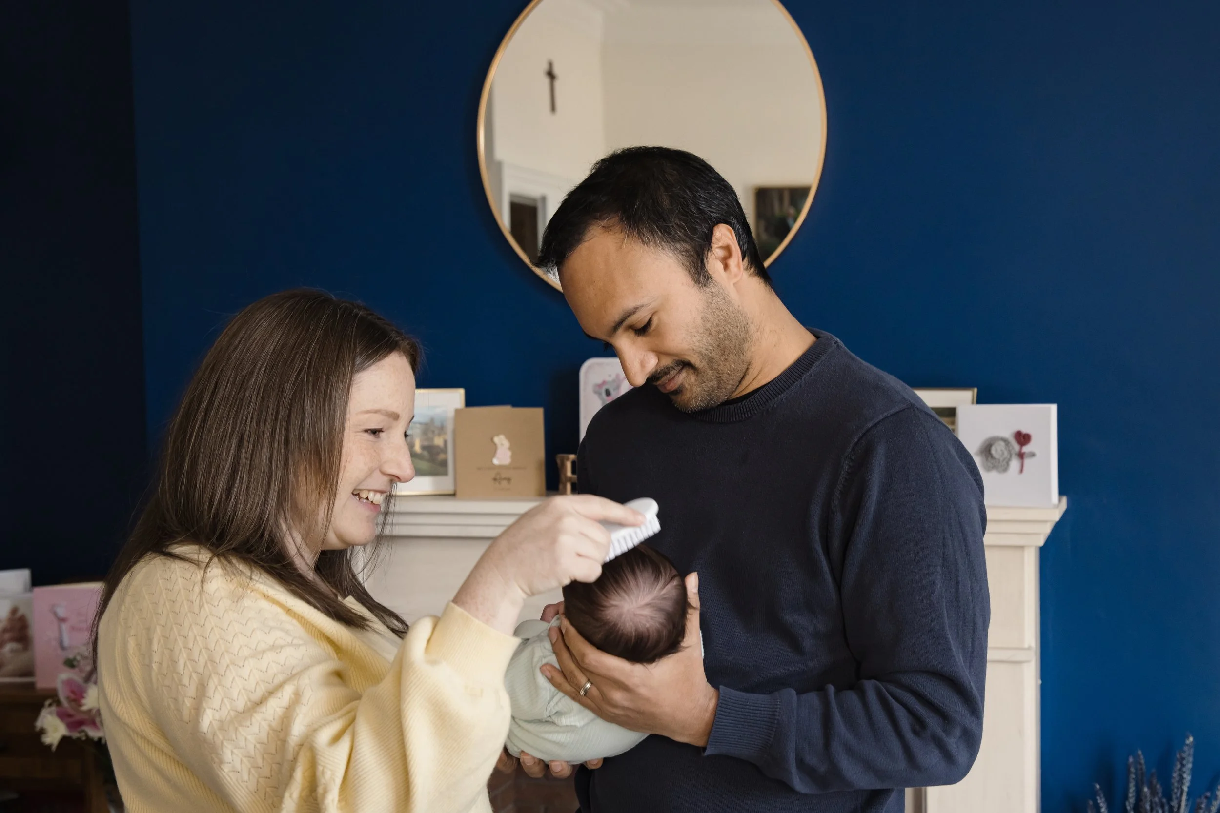 A woman and a man holding a baby, smiling and looking down at the baby, standing in a room with dark blue walls and artwork.