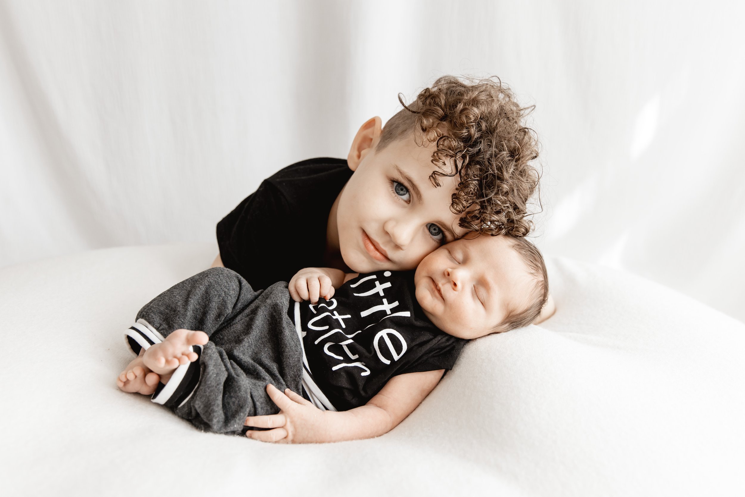 Two young children, one with curly hair and blue eyes and another sleeping baby, lying on a white surface, cuddling together.