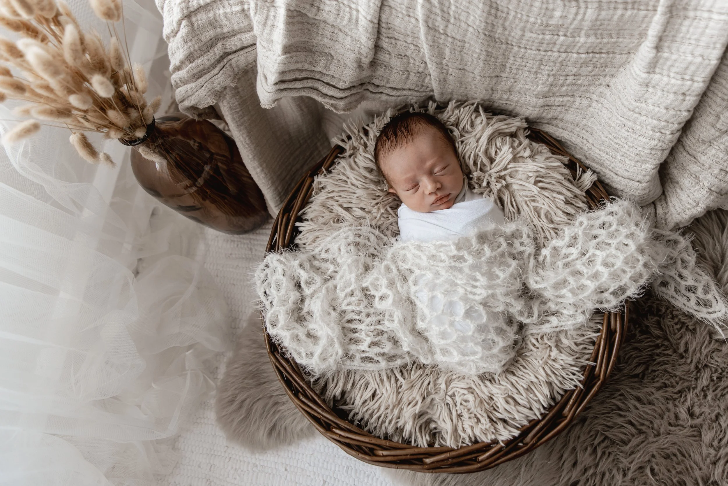 A newborn baby sleeping in a wicker basket, surrounded by cozy blankets and soft textures, with a neutral-colored setting.