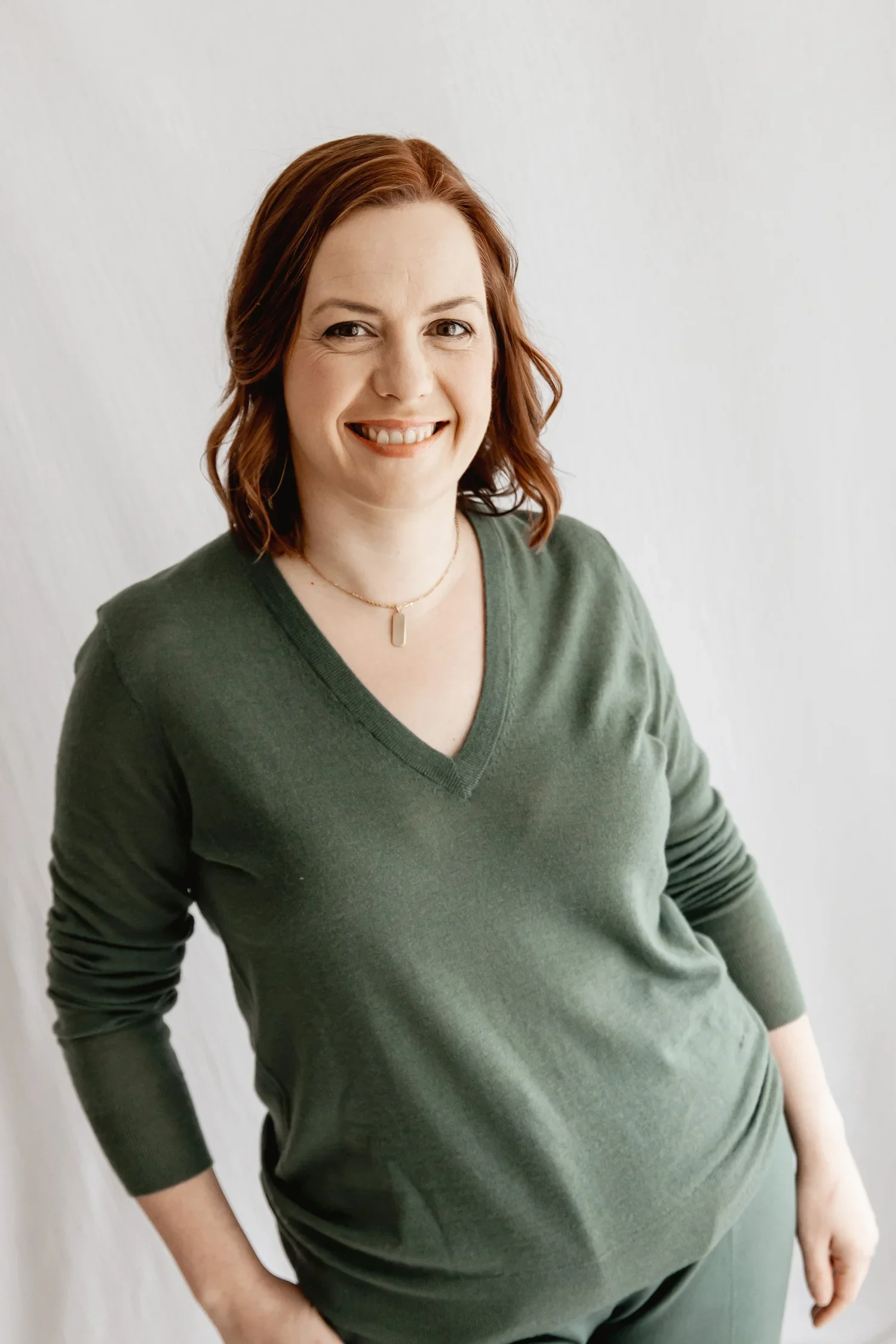 Portrait of a woman with shoulder-length red hair, wearing a green V-neck sweater and a gold necklace, smiling against a plain white background.