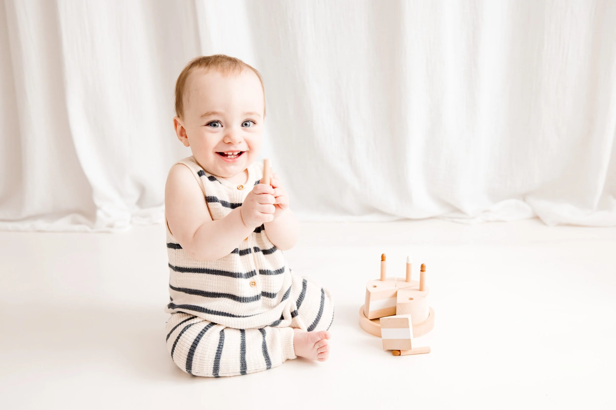 A young child with light brown hair and blue eyes, wearing a striped beige and black sleeveless top, is sitting on the floor with a large cream-colored curtain in the background. The child is smiling while holding a small wooden toy tower, which is p