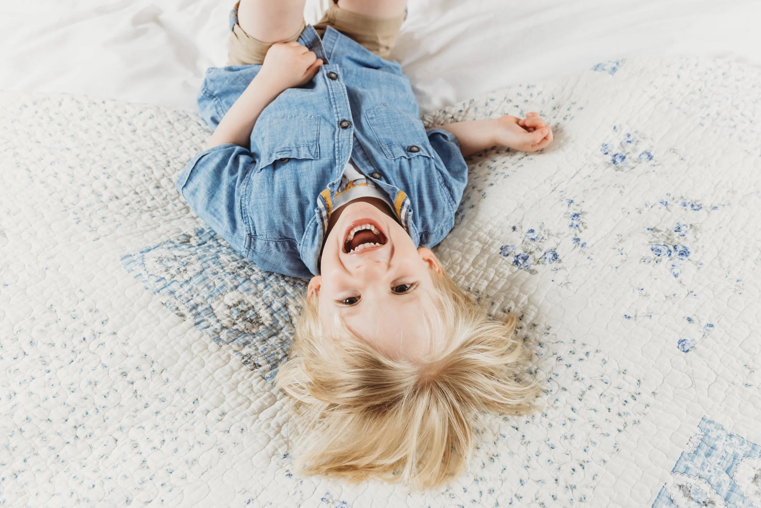 A young child with blonde hair lying on a bed, smiling and wearing a denim shirt.