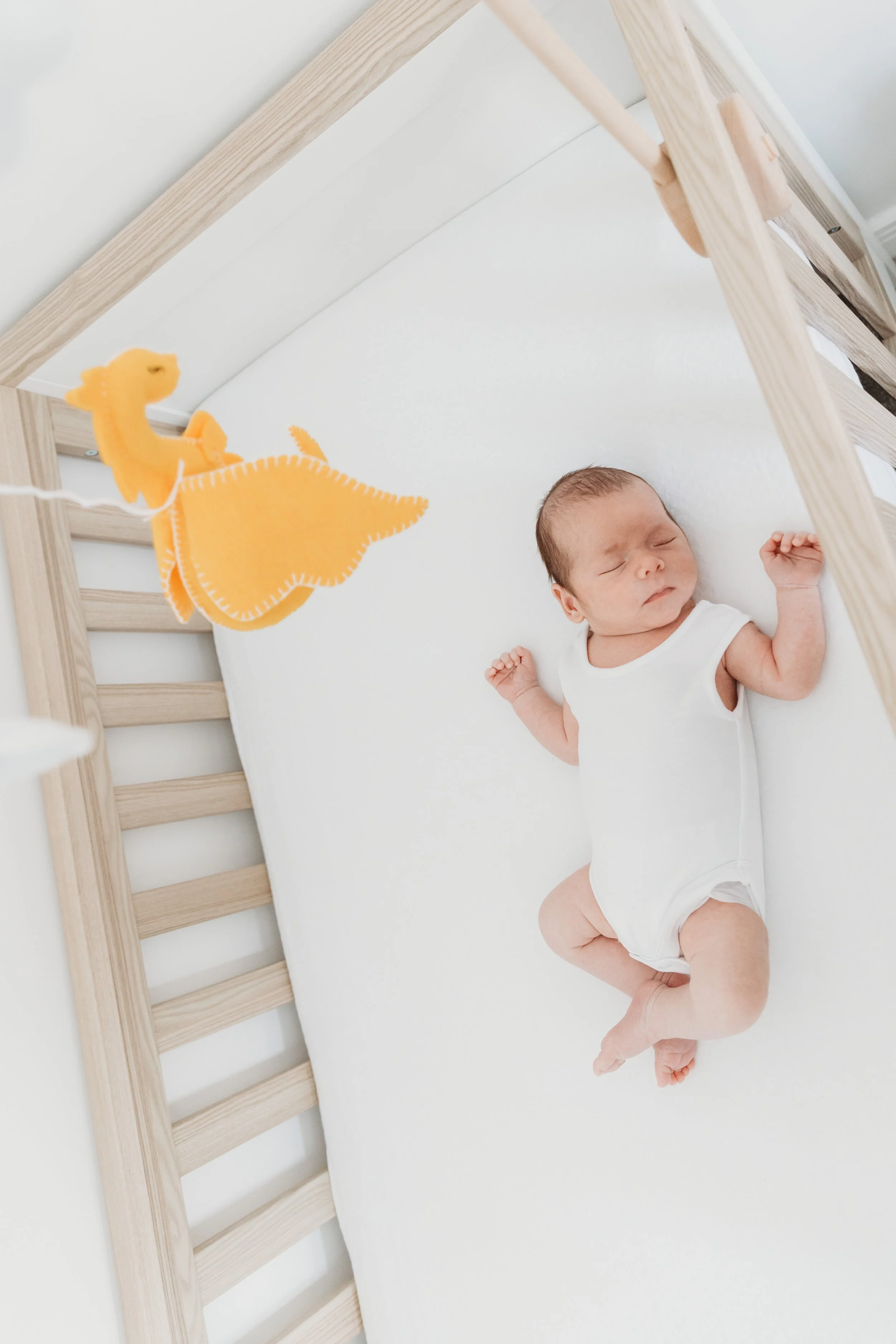 A baby sleeping on a white mattress inside a wooden bed frame with a yellow dinosaur pacifier hanging from the frame.
