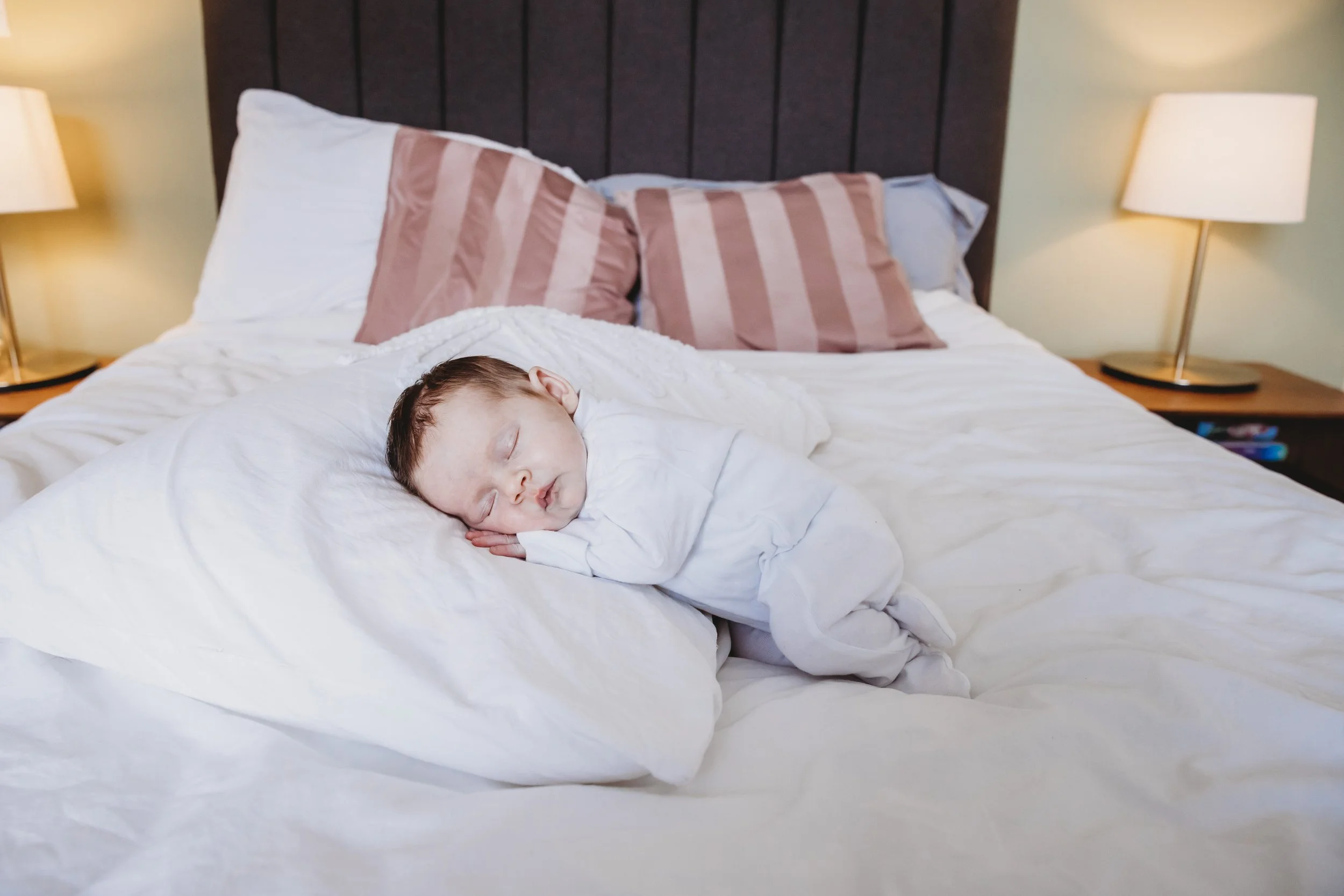 A young boy with light brown hair sleeping peacefully on a bed with white sheets, resting his head on his folded arms. The bed has several pillows and a dark headboard, with two bedside lamps turned on, providing a warm light in the room.