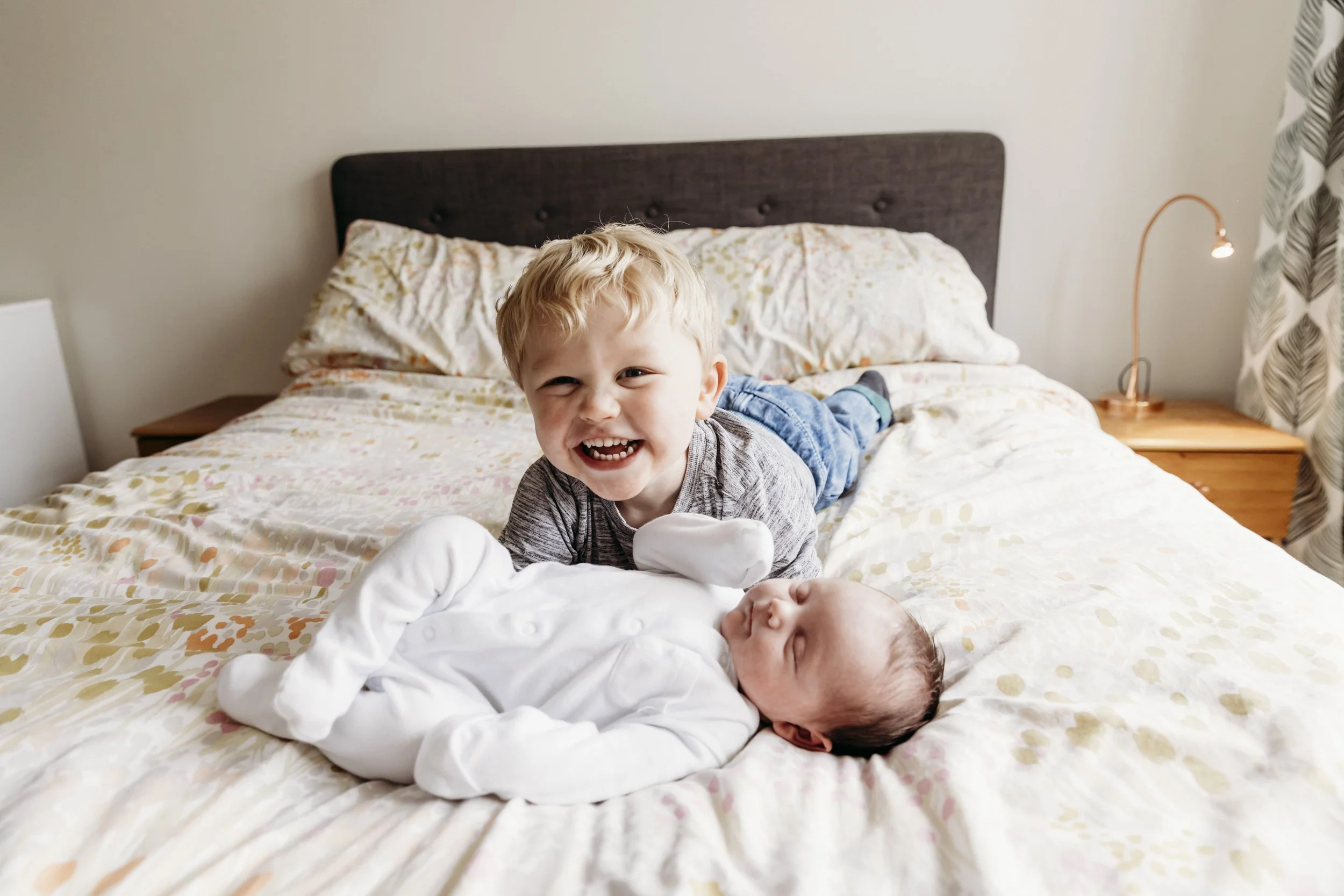 A young boy is smiling and lying on a bed next to a newborn baby, who is sleeping, with both children facing the camera.