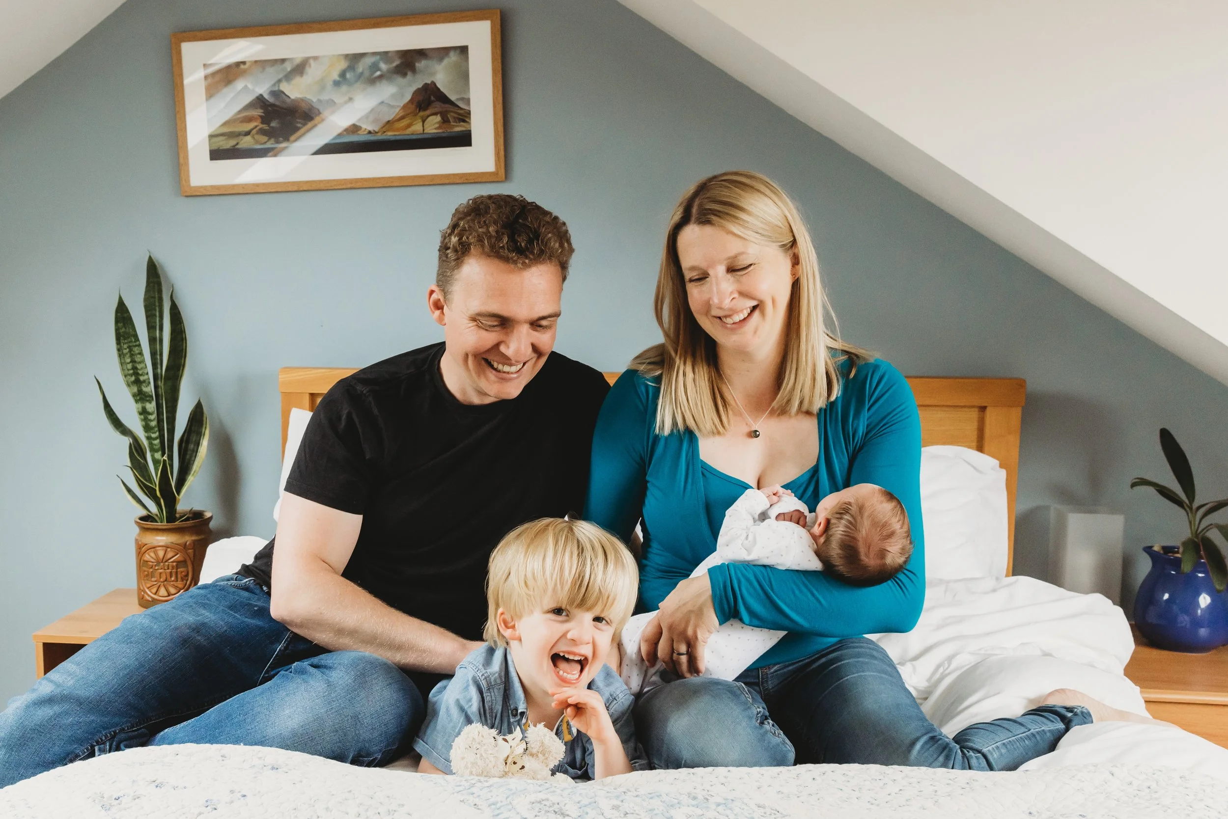 A happy family sitting on a bed in a bedroom, with a woman holding a baby and a young boy laughing, overlooking at one of the children.