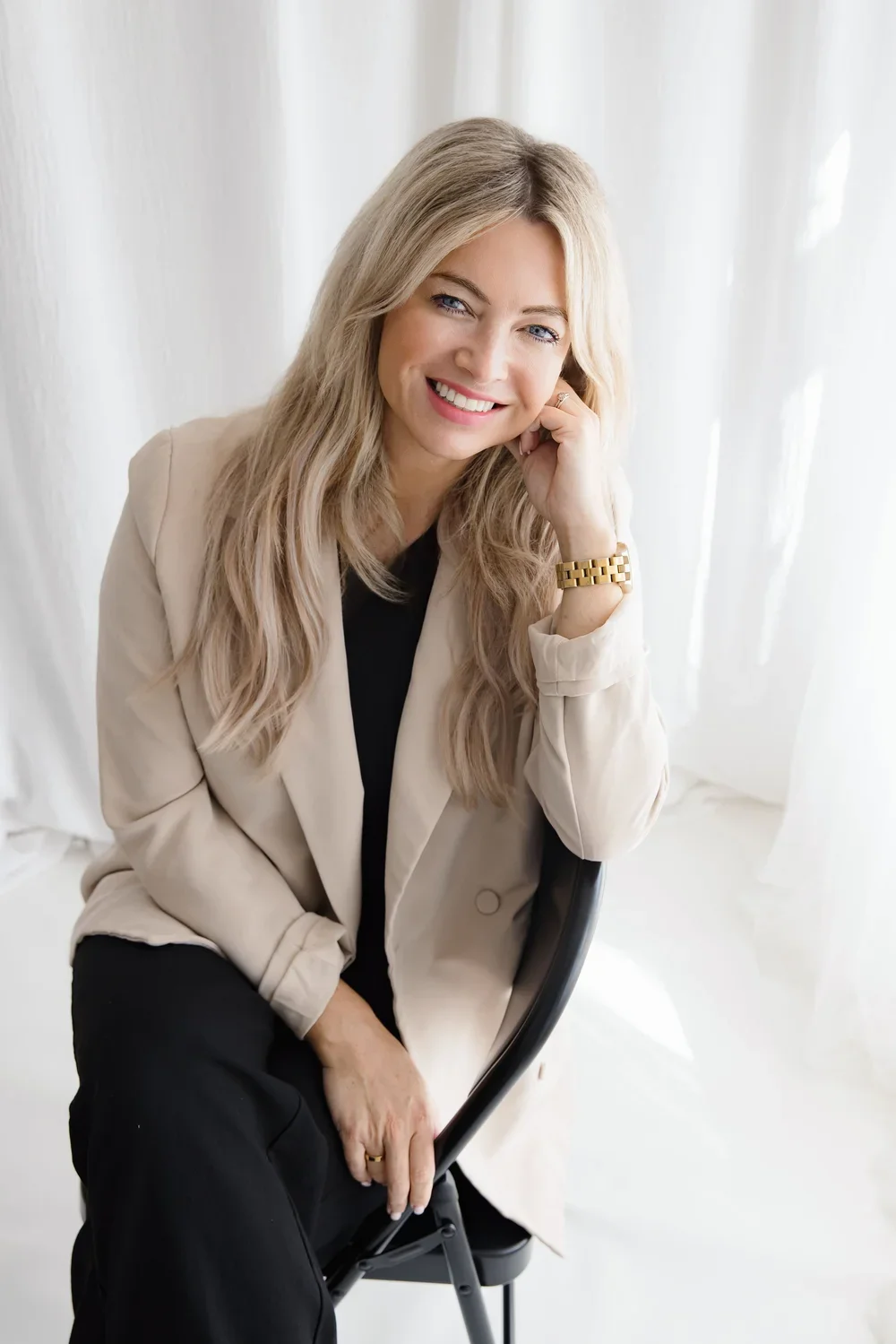 A woman with long blonde hair, sitting on a black chair, smiling with her head resting on her hand, wearing a beige jacket, black top, black pants, and a gold watch.