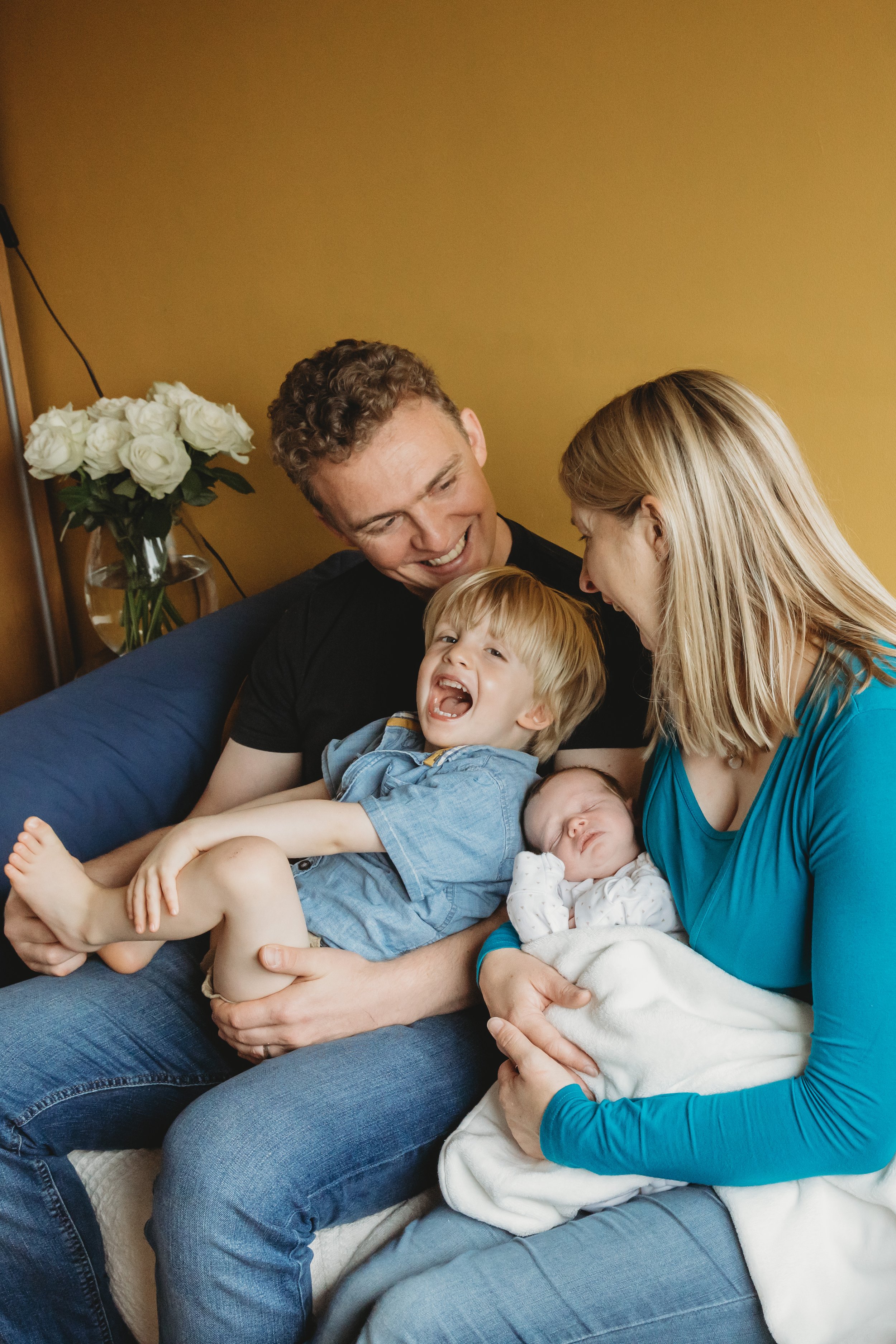 Family sitting on the sofa, smiling, and playing with their children in a cozy living room with a yellow wall, a vase of white roses, and a blue sofa.
