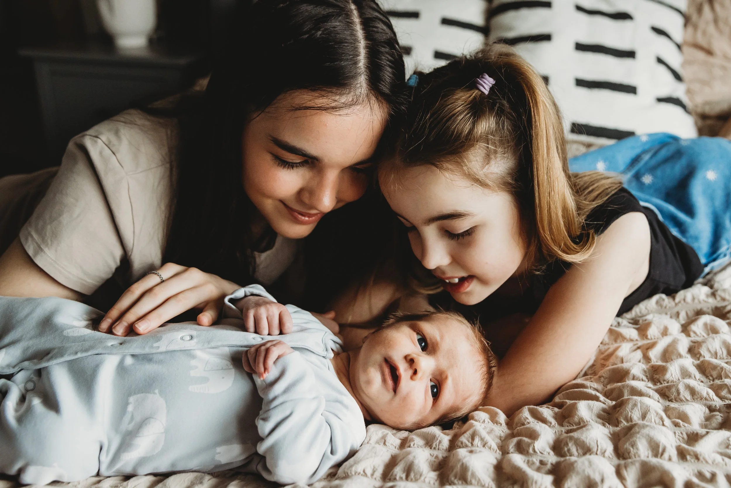 A woman and a young girl are lying close together on a bed, looking at a baby lying on its back. The woman and girl have happy, affectionate expressions as they gaze at the baby, who is wearing a onesie and looking up.