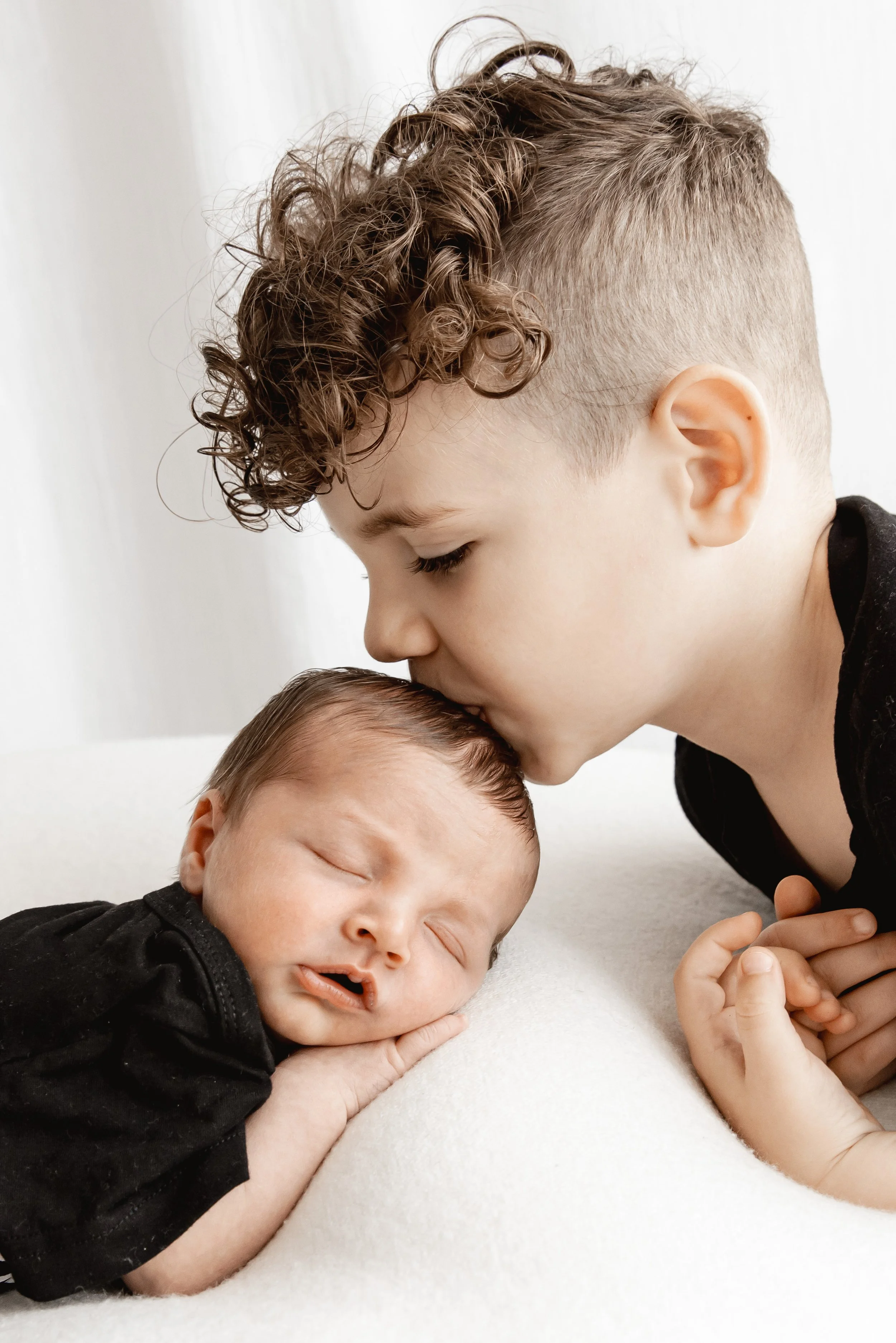 A young boy with curly hair leaning over a newborn baby sleeping on a white surface, with his face close to the baby and eyes closed.