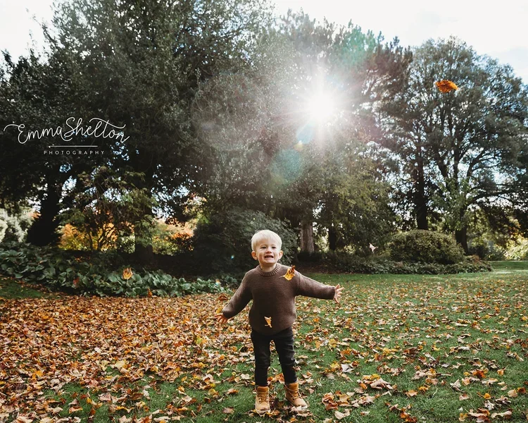 A young child with blonde hair standing in a park surrounded by fallen autumn leaves, smiling and playing with falling leaves as the sun shines brightly through the trees.
