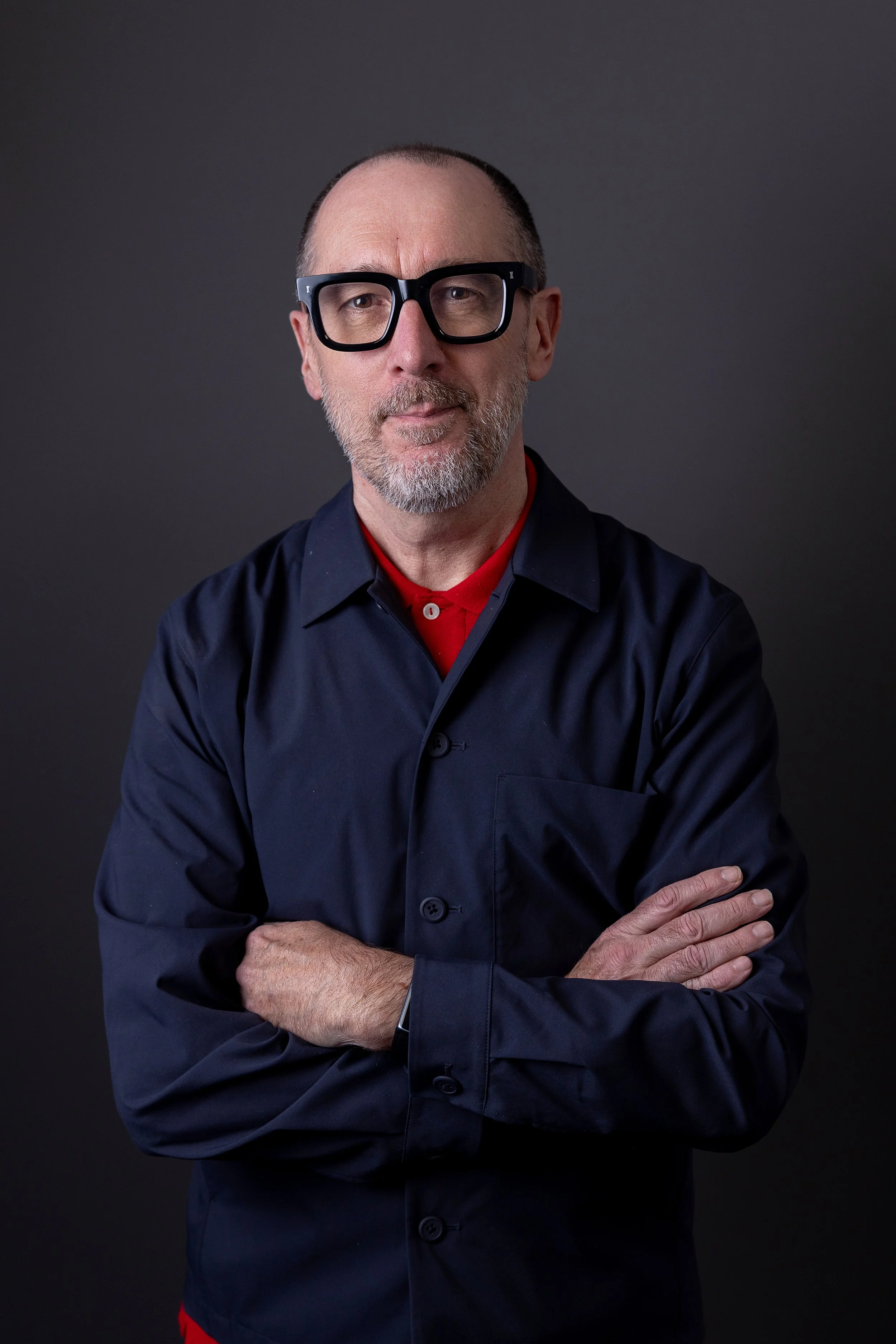 Portrait of a middle-aged man with a beard and glasses, wearing a black jacket and a red shirt, posing with arms crossed against a dark background.