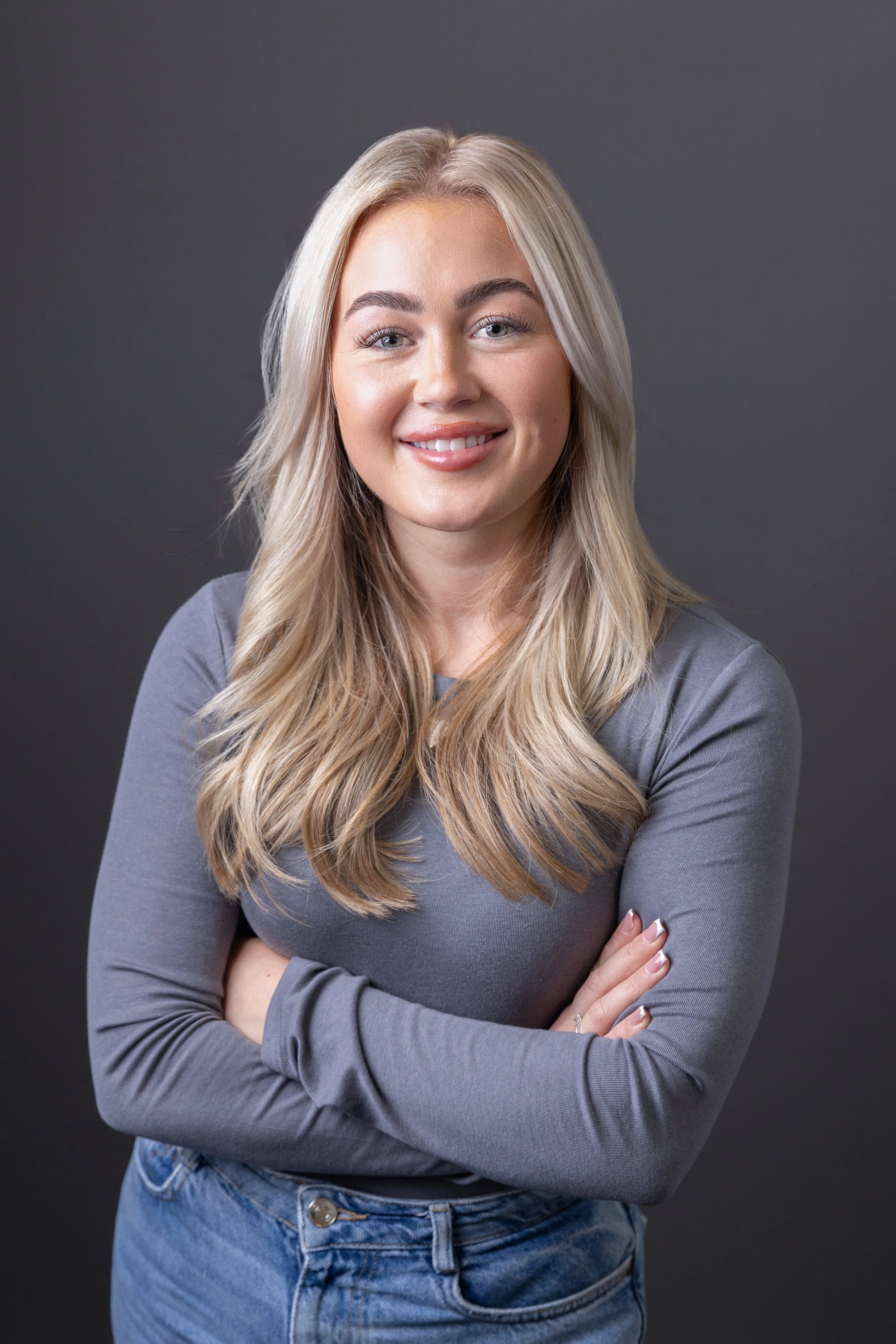 A young woman with long blonde hair and light skin, wearing a long-sleeve gray top and blue jeans, smiling with her arms crossed against a dark gray background.