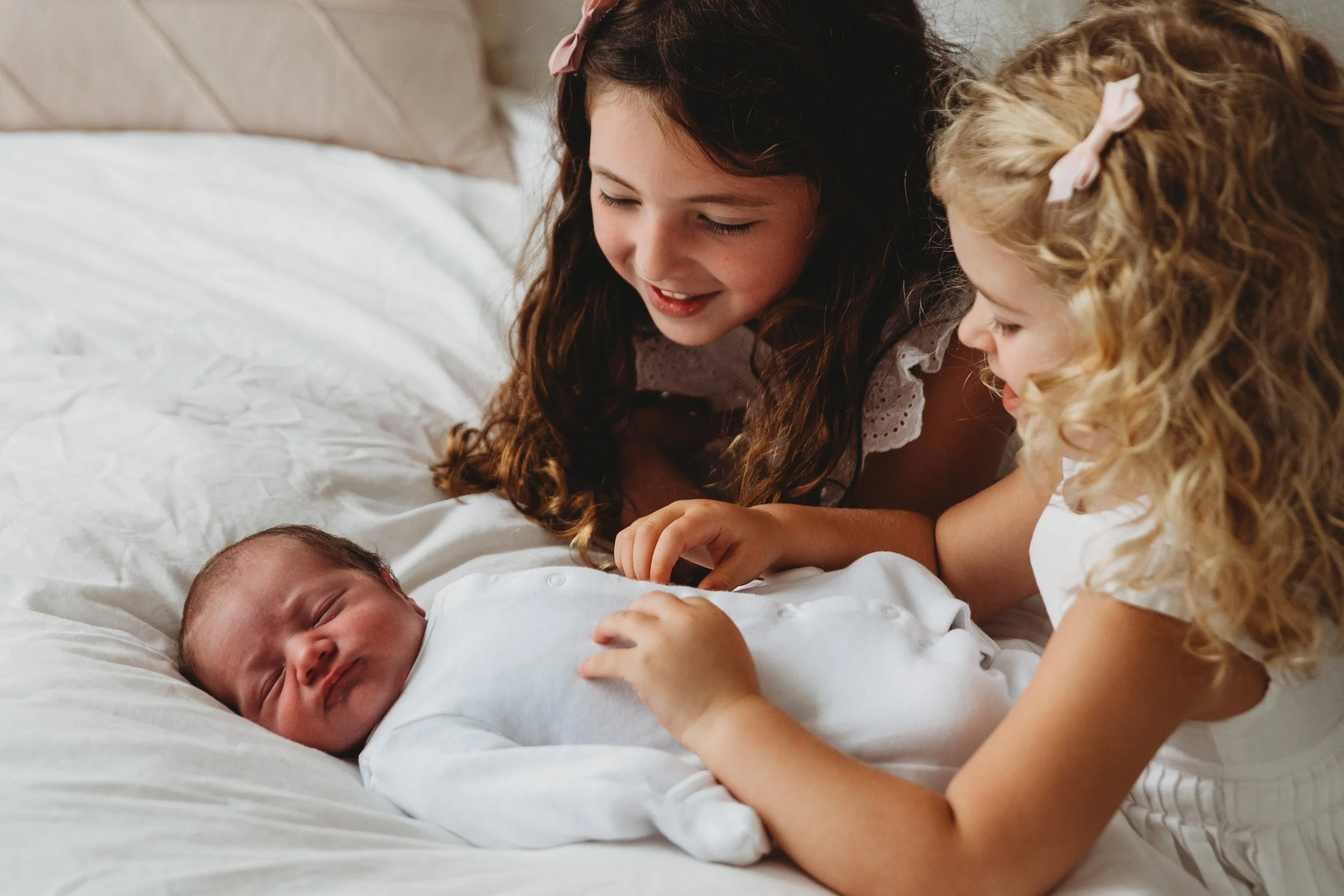 Three young girls with long hair, wearing pink and white bows, looking down and smiling at a newborn baby lying on a bed. The baby is dressed in white and appears to be sleeping.