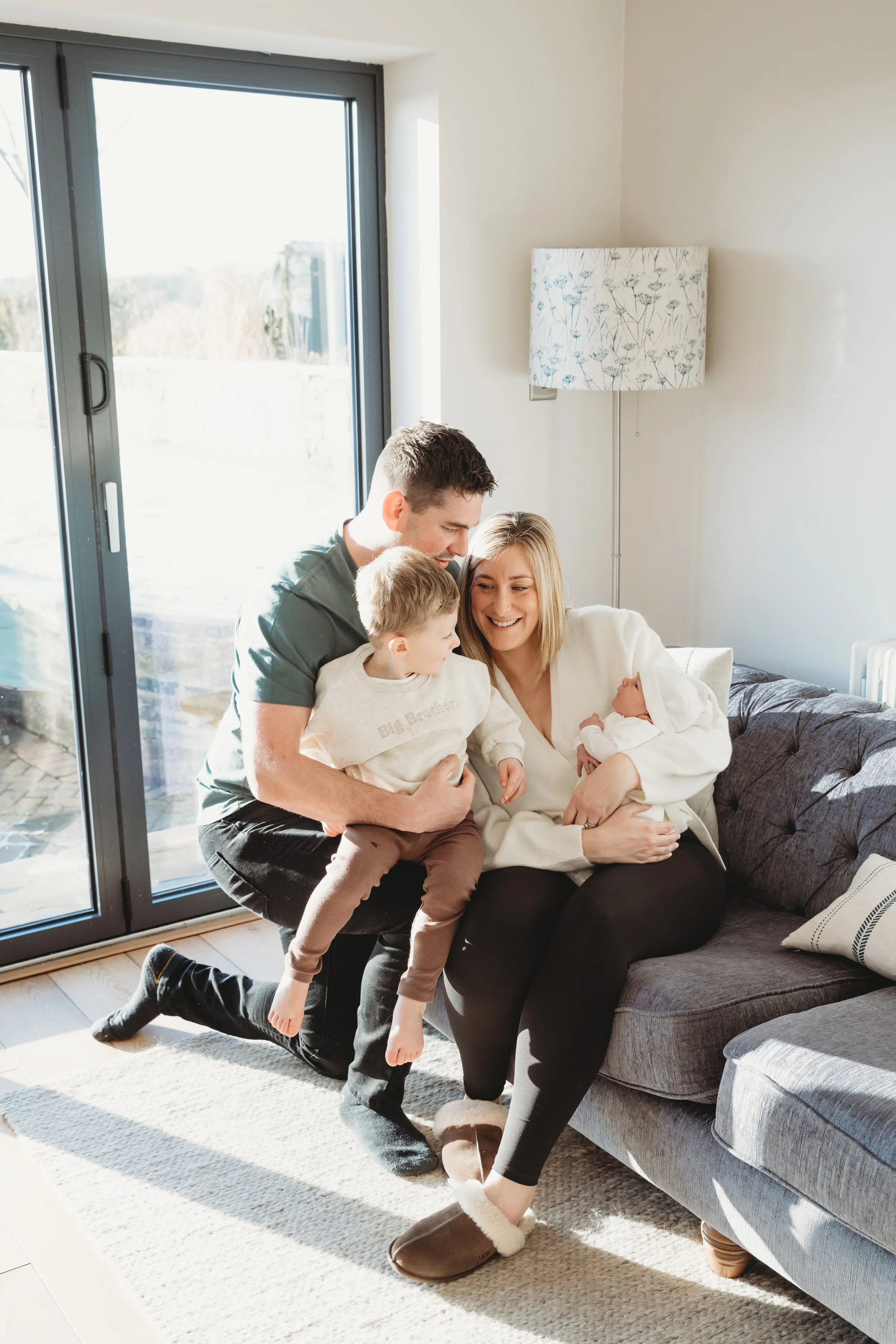 A family of four sitting together in a bright living room, smiling and enjoying time with their two children, one of whom is a baby in a white outfit.