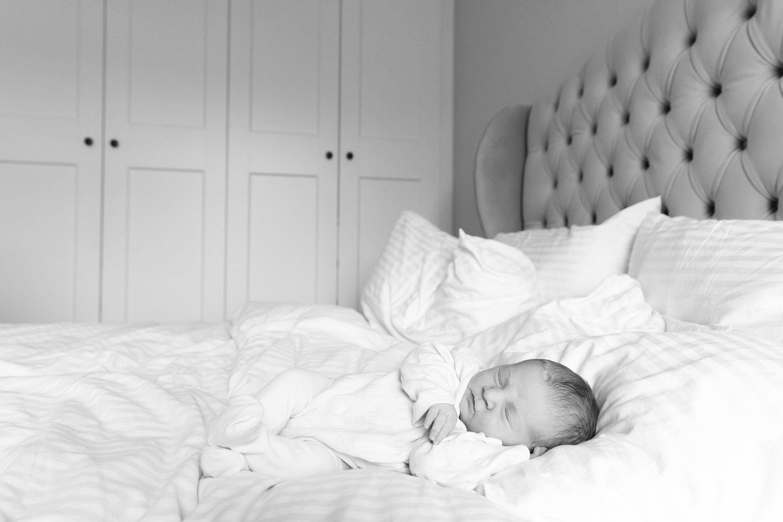 A newborn baby sleeping on a bed with white bedding, in front of a tufted headboard, with a clothes closet with closed doors in the background.