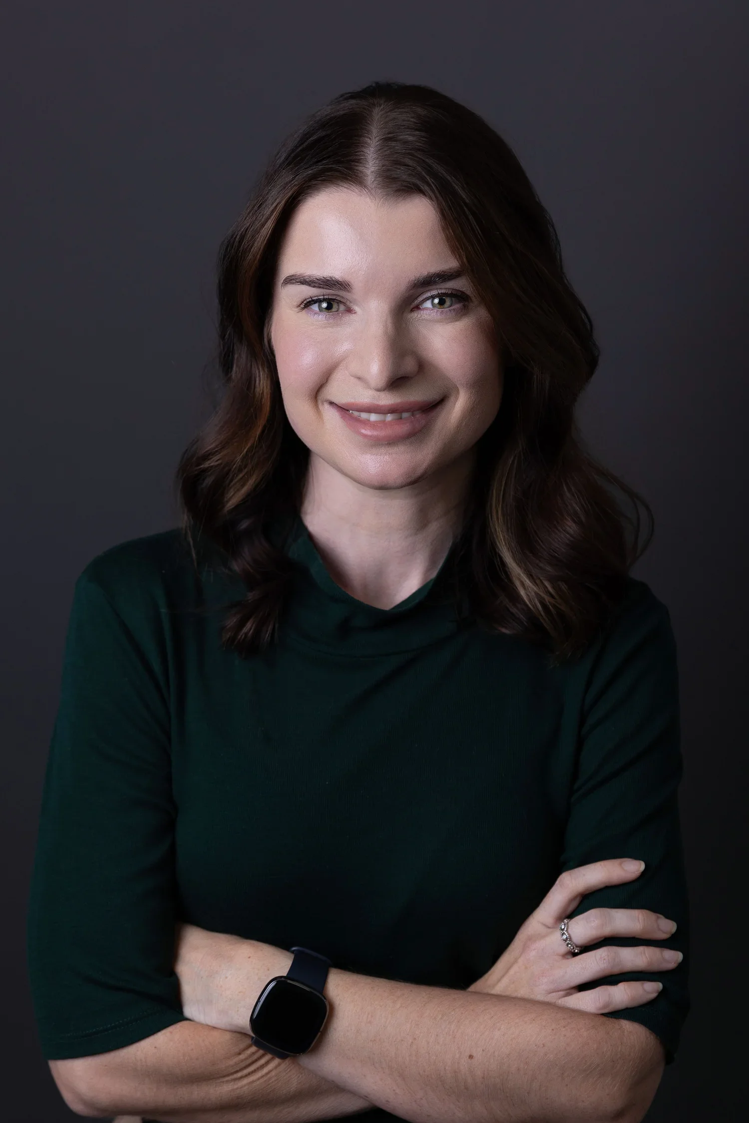 Portrait of a smiling woman with medium-length wavy brown hair, crossing her arms, wearing a dark green top, black smartwatch, and a ring, against a dark background.